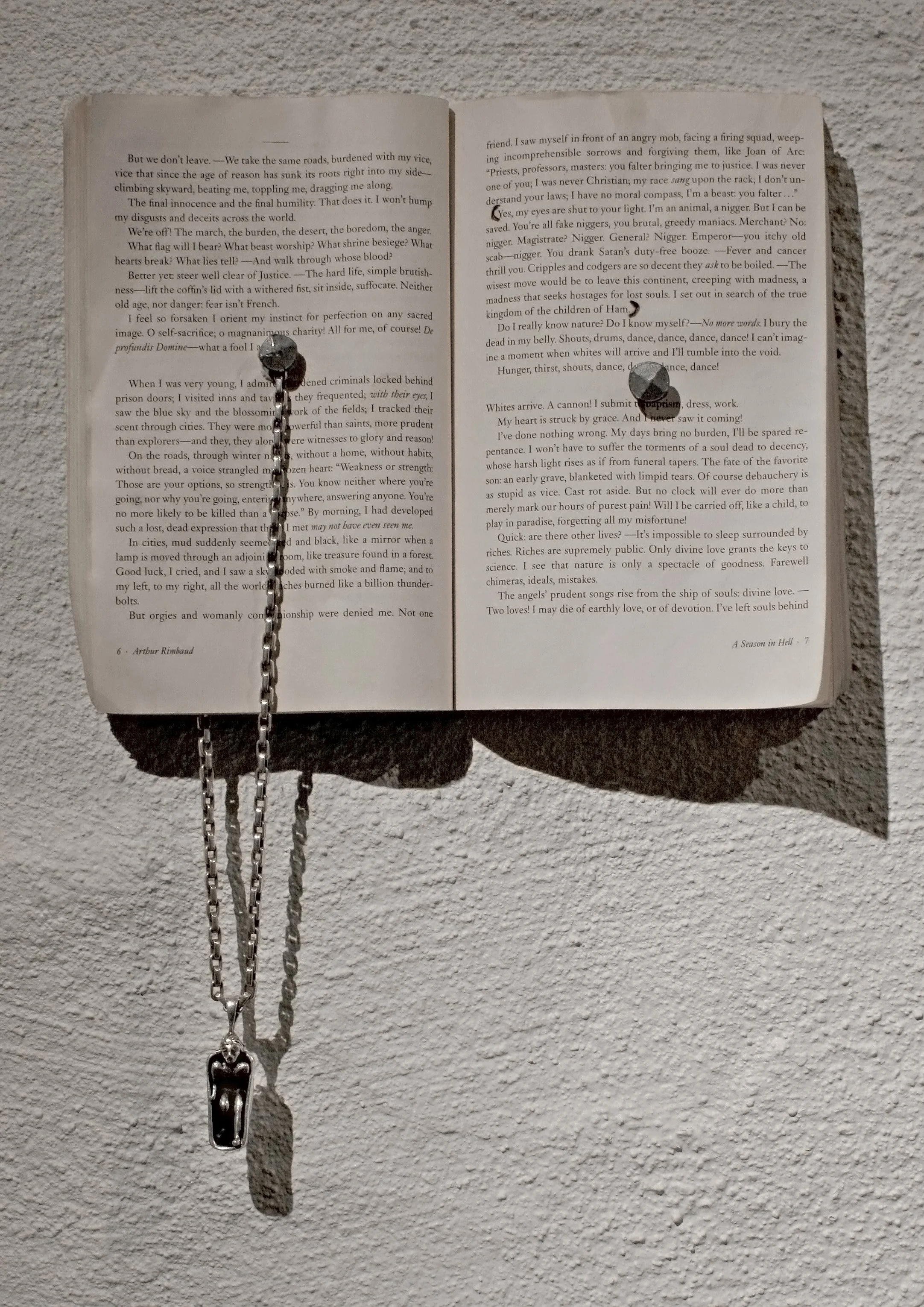 An open book on a textured surface with a chain and a pendant hanging from the left page, Jim Morrison replacing Jesus Christ and the bath tub replacing the cross, casting shadows. The book A Season in Hell by Rimbaud.