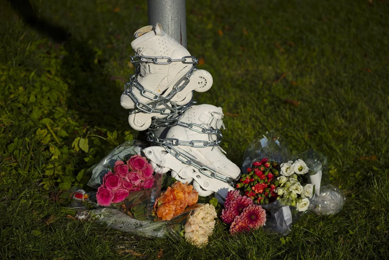 A pair of white roller skates chained to a pole, surrounded by colorful flowers with some wrapped in plastic. Referring to ghost bikes, but these are ghost blades, relating to the speed crisis in Oslo