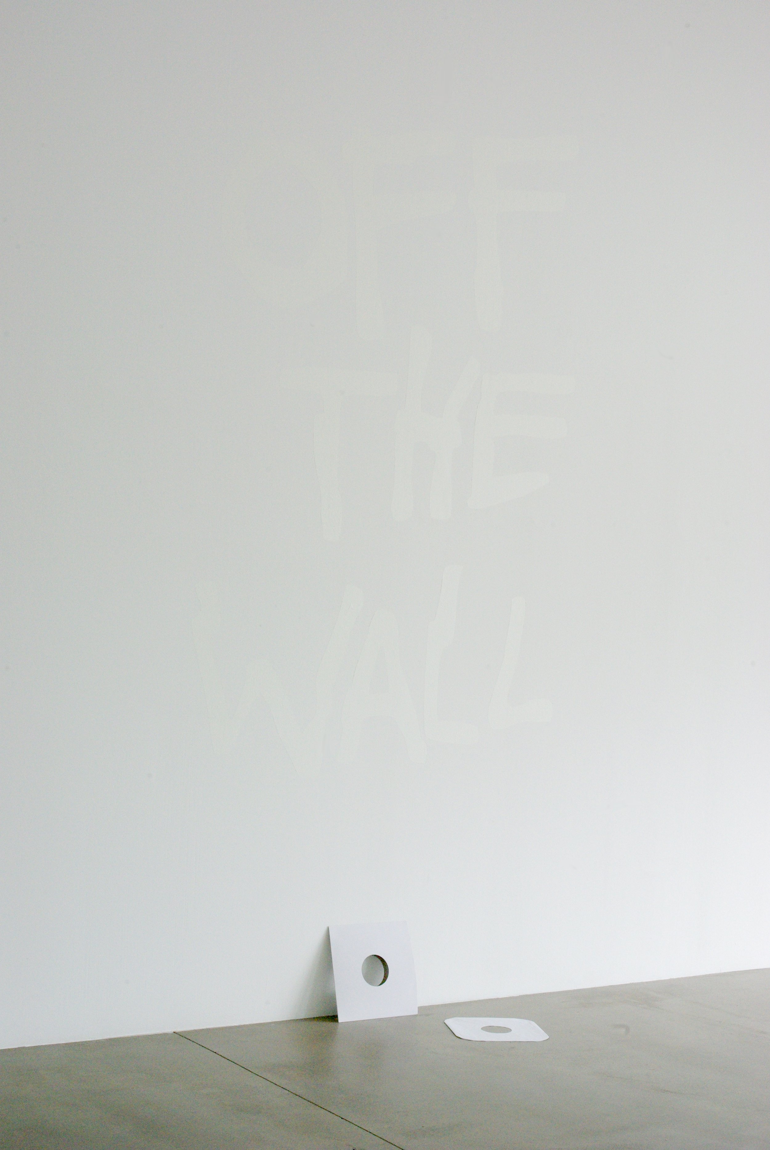 Gallery wall with two square white frames featuring circular cutouts, leaning on a gray concrete floor against a plain white wall. Invisible ink says OFF THE WALL.