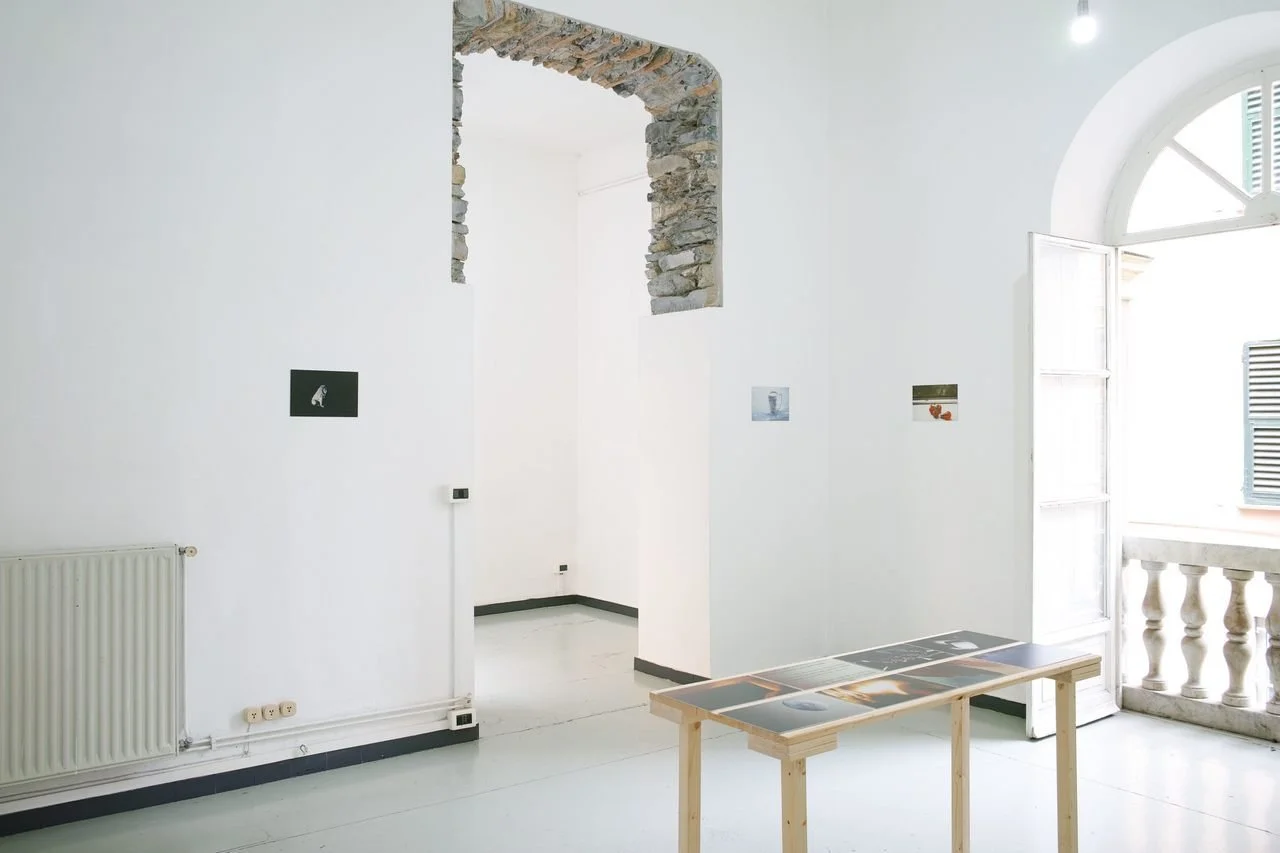 An art gallery with white walls, a small table with photographs, small images on the walls, a stone archway, and an open balcony door with balusters. Space 4235, Genova.