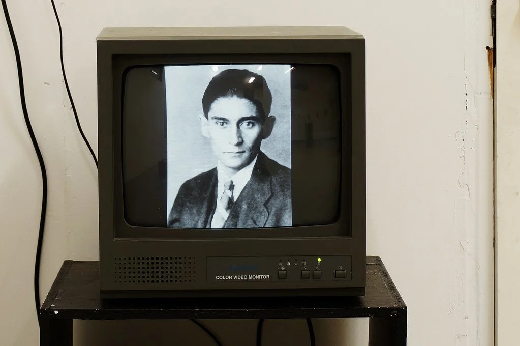 A vintage black-and-white photograph of a young man in a suit is displayed on a small CRT television on a black table, against a plain white wall.