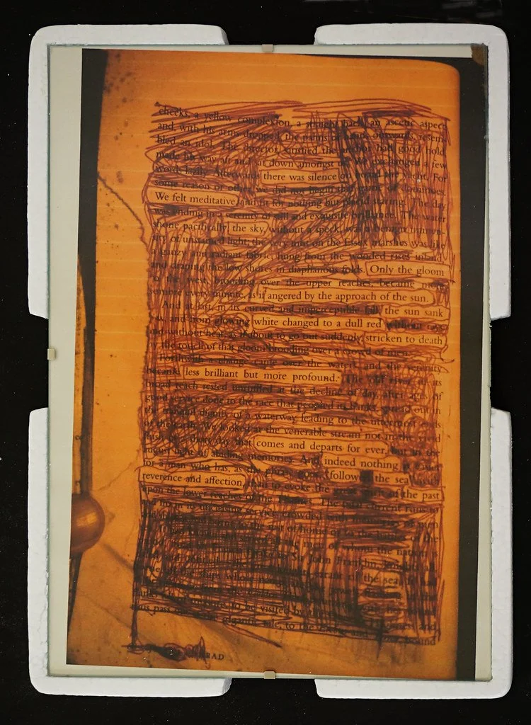 A close-up photograph of an orange-colored page with typed text, heavily scribbled over with black ink, making the text difficult to read.