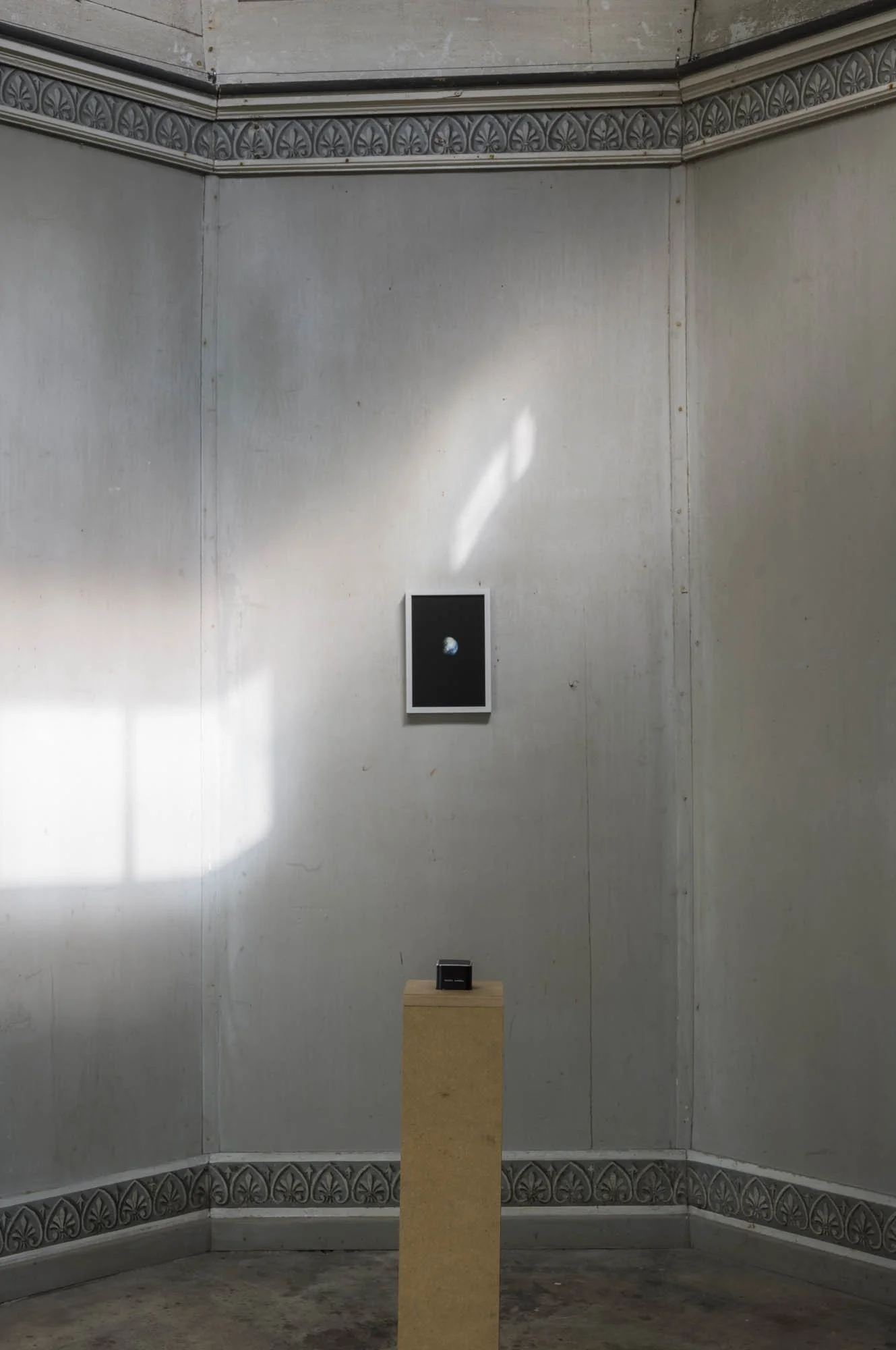 An art installation in a corner of a room with two artworks displayed on the wall, one framed picture and one small object on a pedestal, with sunlight casting shadows on the wall.