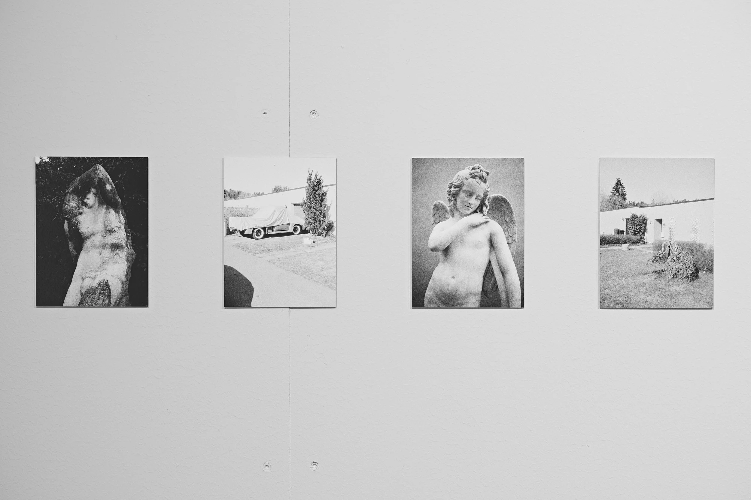 Four black and white photographs displayed on a gallery wall. The first shows a carved wooden sculpture of a horse, the second a parked car covered with a cloth next to a tree, the third a sculpture of a cherub with wings, and the fourth a yard with 