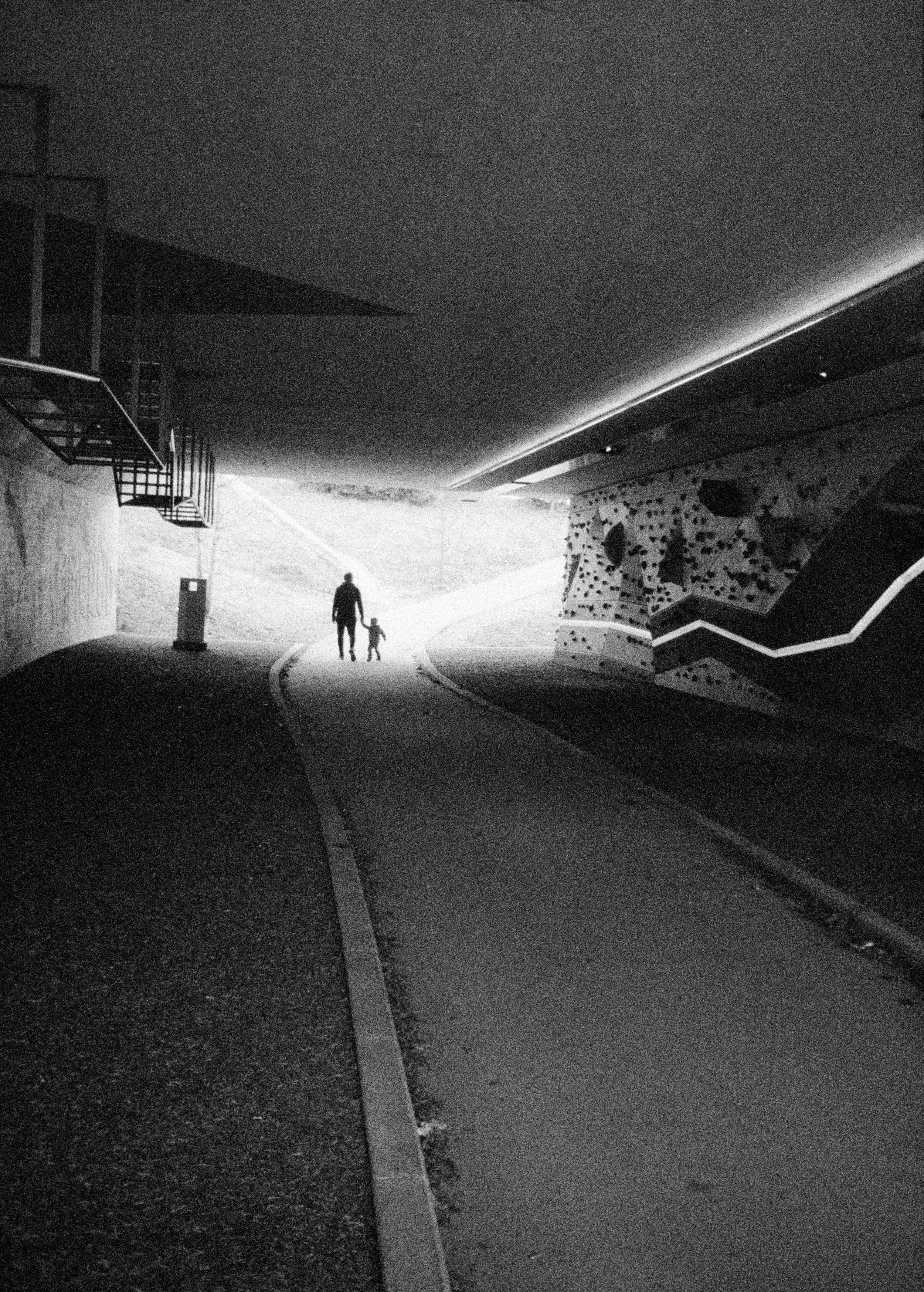 Silhouette of a person and a child holding hands, walking through a tunnel towards bright light outside. Ilford Delta 3200. Olympus half frame camera.