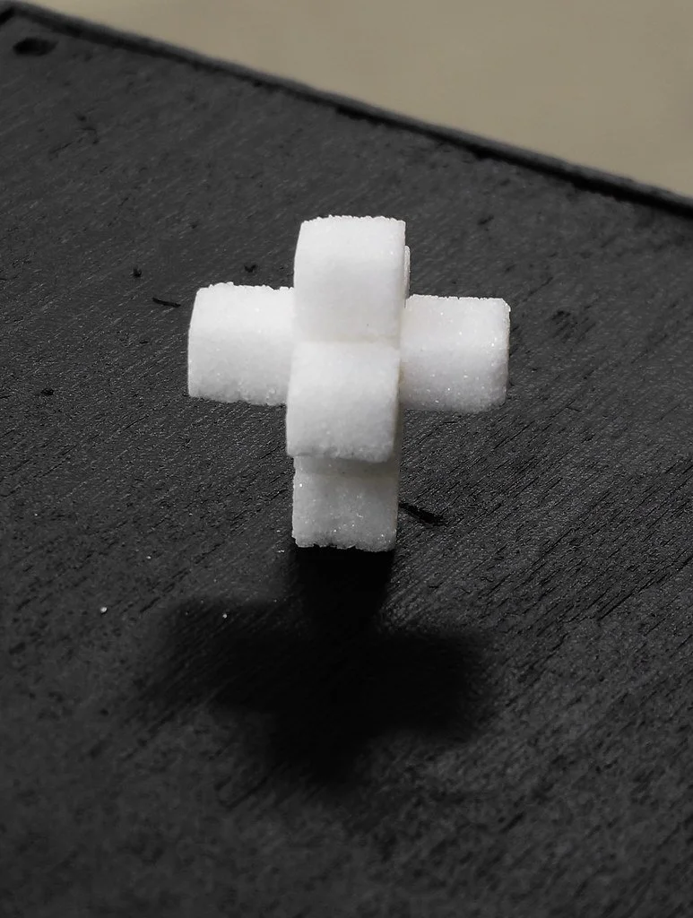 Six white sugar cubes arranged in the shape of a plus sign on a dark surface with a shadow cast underneath.