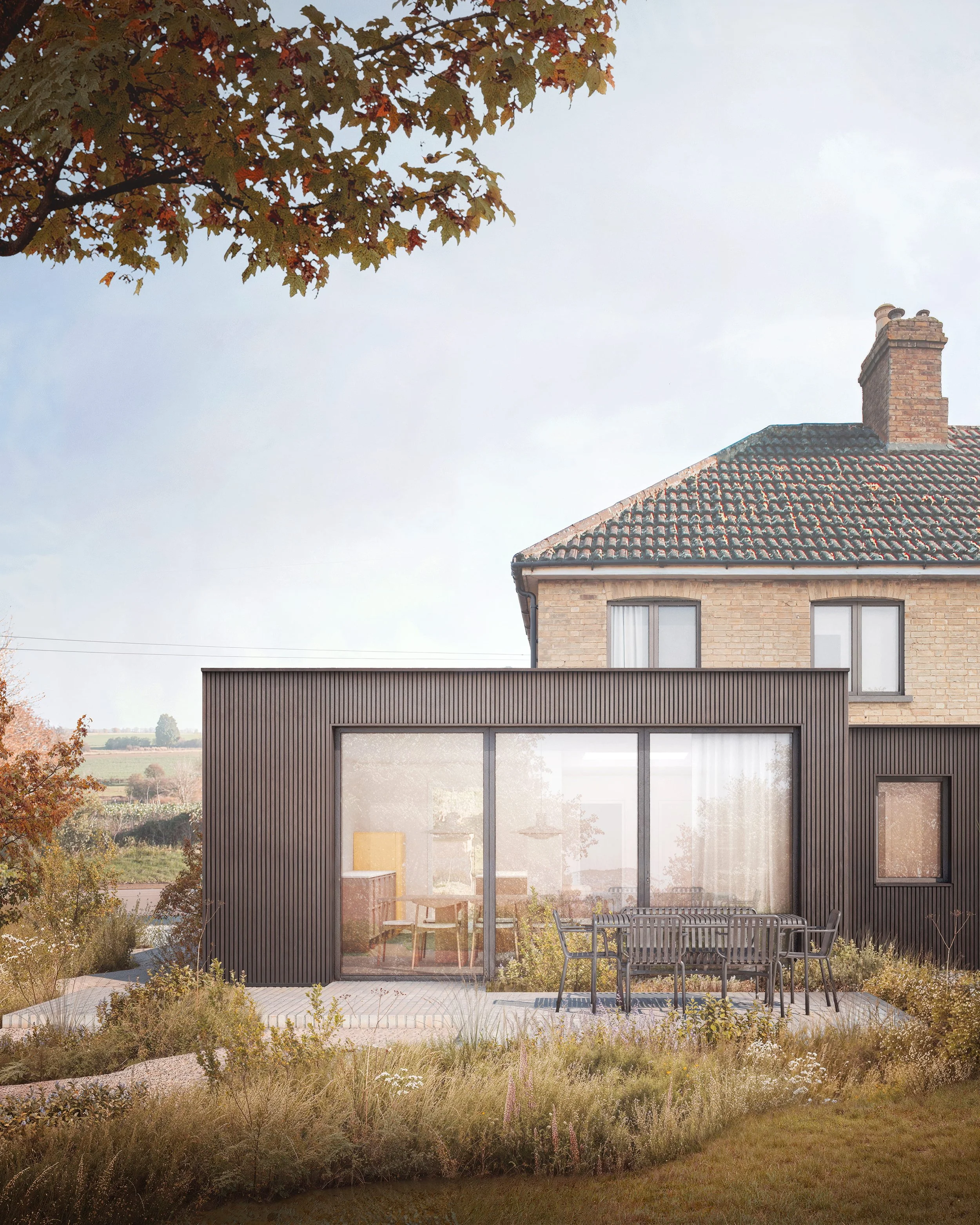A modern extension with a black timber exterior and large glass doors attached to a traditional brick house with a tiled roof. There is outdoor dining furniture on a patio surrounded by a garden with various plants and trees.