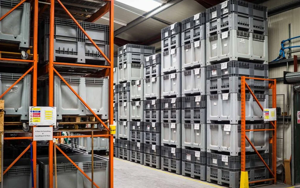 Stacks of large gray plastic storage crates in a warehouse, organized on orange metal shelving units.