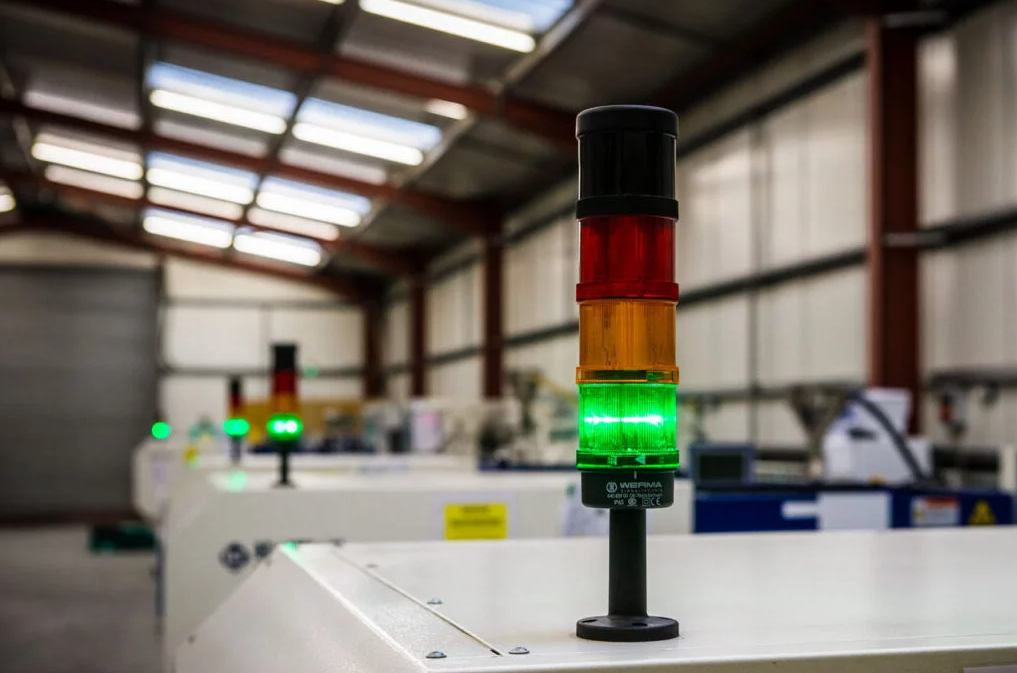 Industrial warning light system with three colored lights — black at the top, red, yellow, and green underneath — in a manufacturing or warehouse setting.