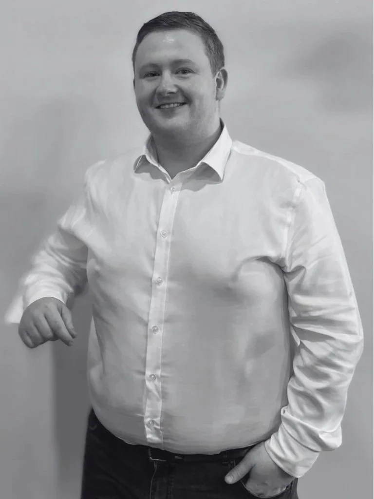 A smiling man with short hair wearing a white button-up shirt and dark pants, standing against a plain background.