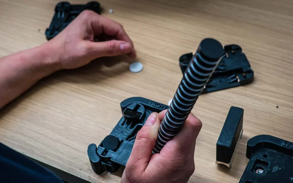 A person assembling or repairing a firearm, with the slide and other parts on a wooden surface. The person is holding a coil spring, part of a gun recoil assembly.