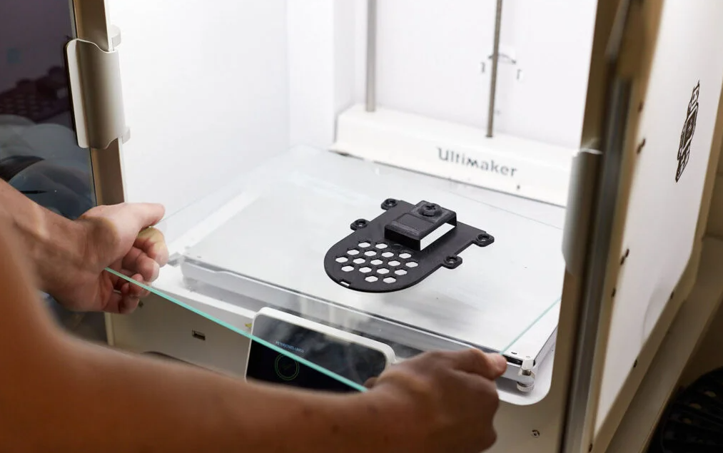 Person holding a glass door while inspecting a transparent print bed with a 3D printer inside an Ultimaker machine. A finger is pushing against the glass surface near the edge.
