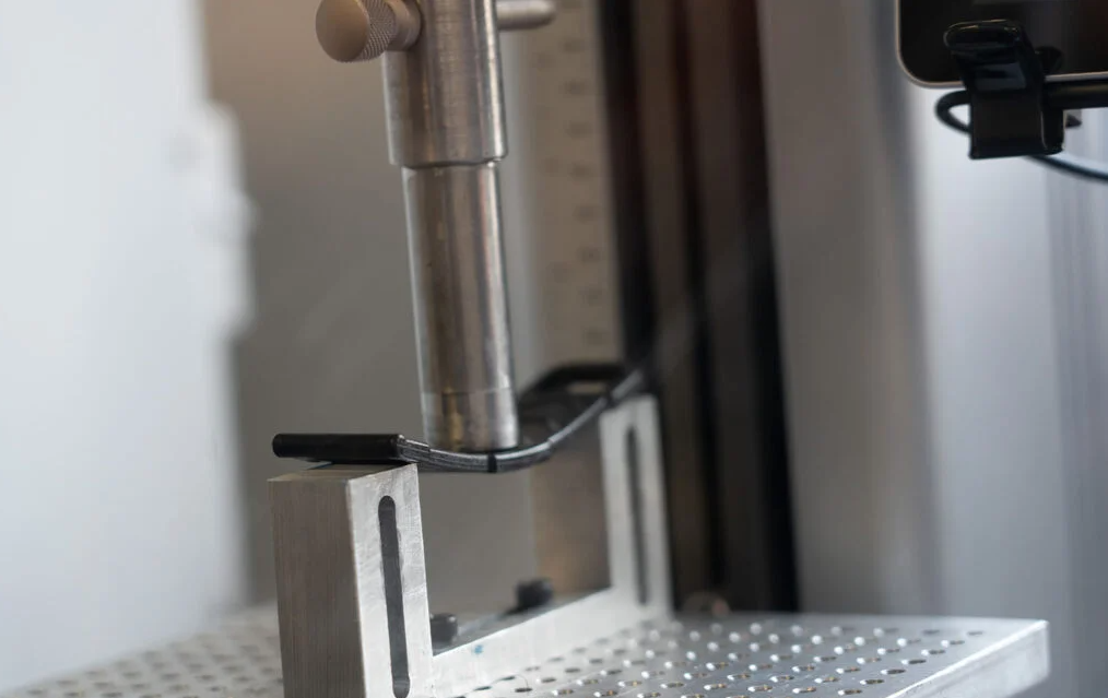 Close-up of a metal stand holding a long cylindrical metal tool or instrument, with a black cable running along a metal frame, in a laboratory or industrial setting.