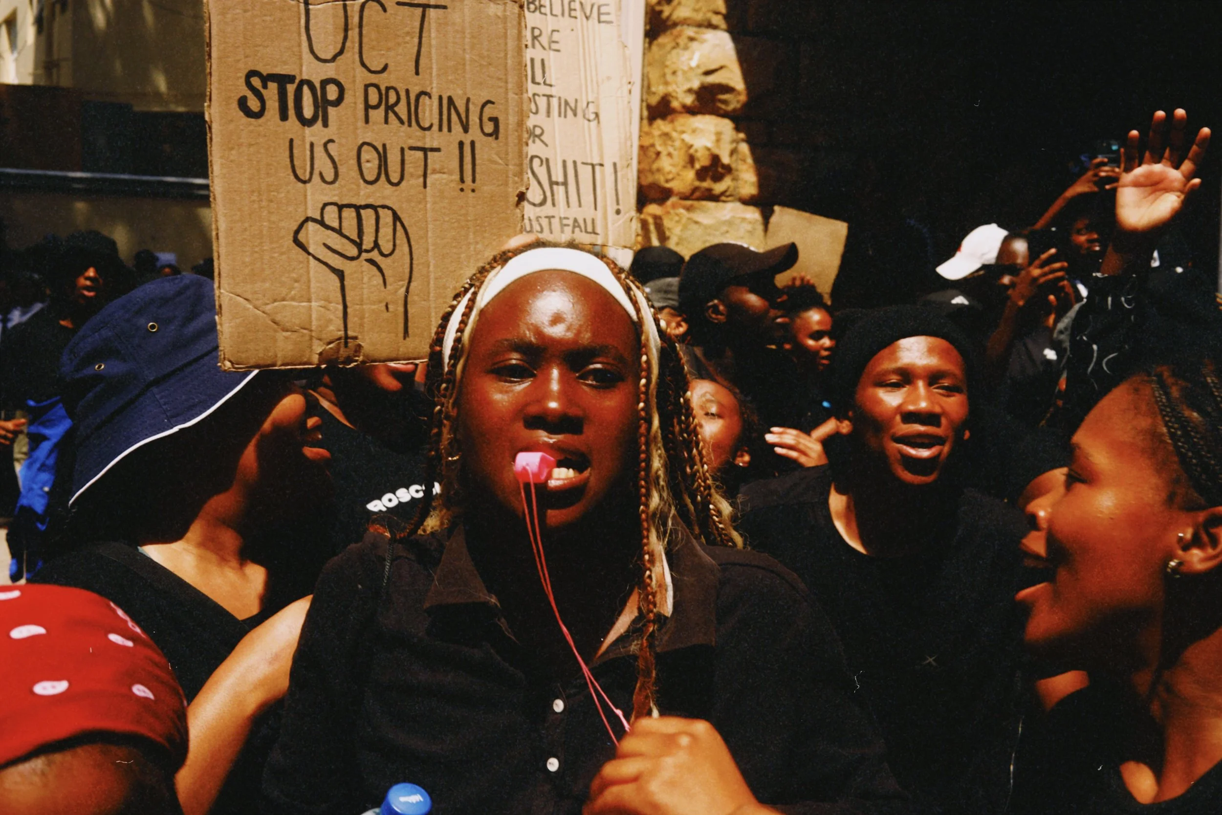 A group of protestors, predominantly women, gathered at night, with one woman holding a cardboard sign that reads 'STOP PRICING US OUT!!' and raising a fist symbol. The woman in the center has braided hair, appears to be speaking into a microphone or headset, and is surrounded by other women expressing strong emotions.