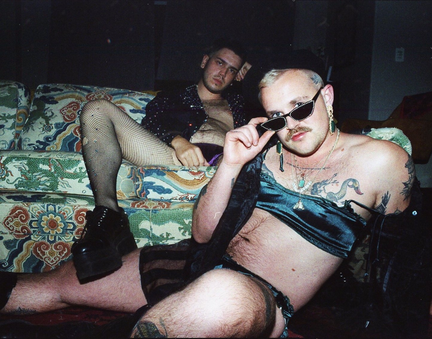 Two men posing on a colorful vintage couch in a casual indoor setting, one wearing sunglasses and jewelry, the other in fishnet stockings and boots.