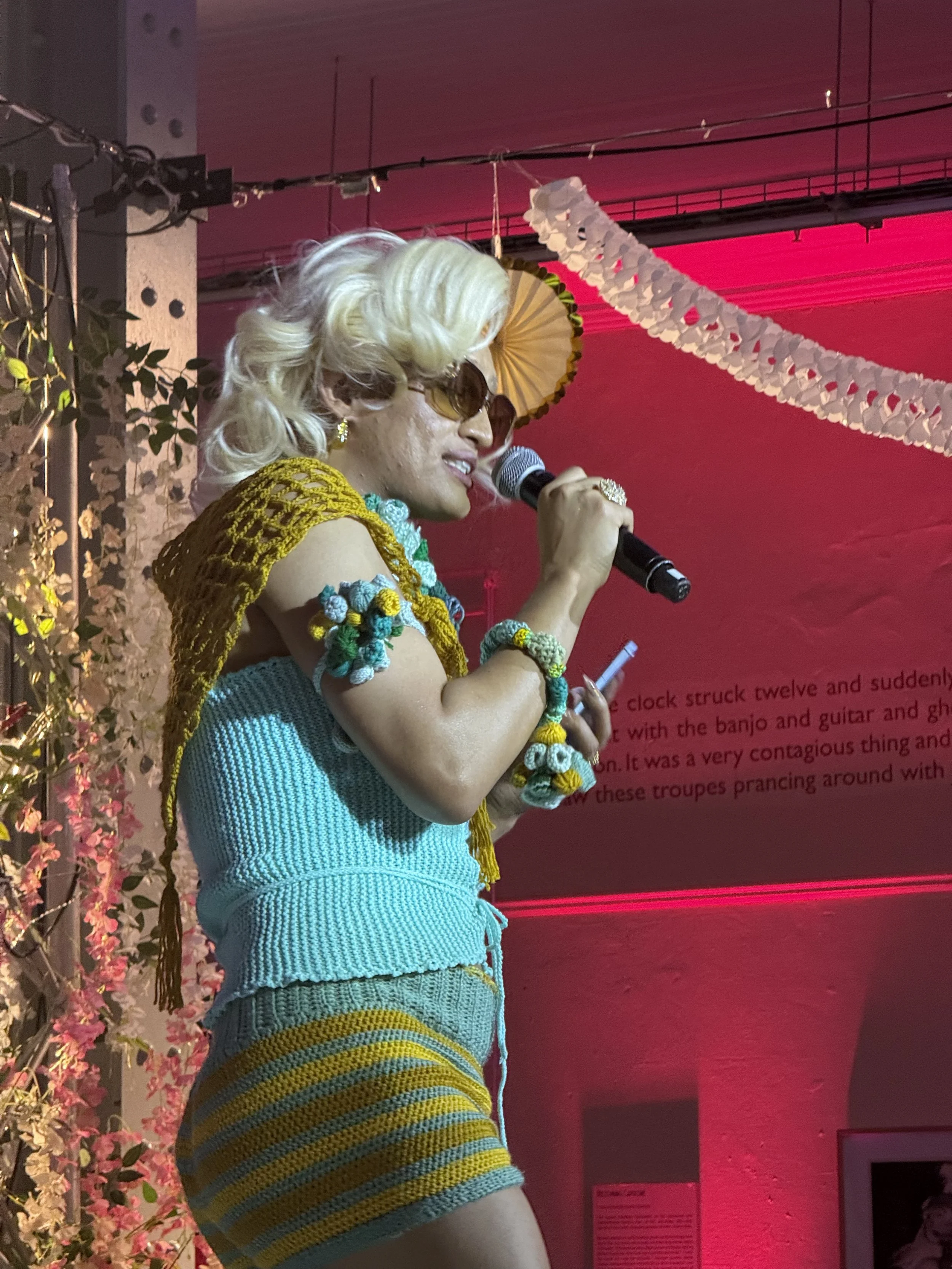 Person dressed in colorful knitted outfit, with blonde wig and sunglasses, speaking into a microphone at a decorated indoor event.