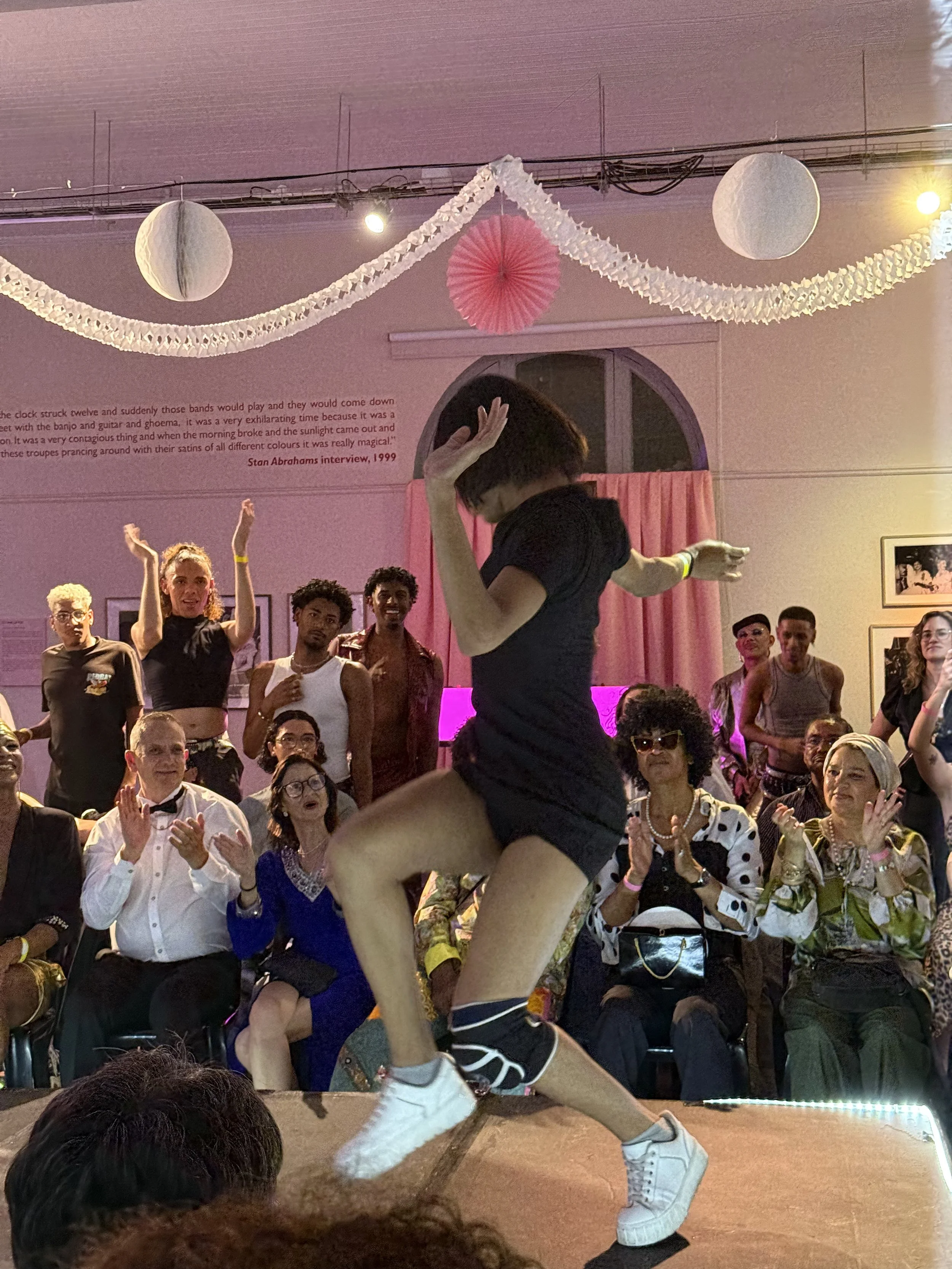 A woman dancing on stage with a crowd of people clapping and watching her. The setting appears to be a lively party or performance event with decorations hanging from the ceiling.