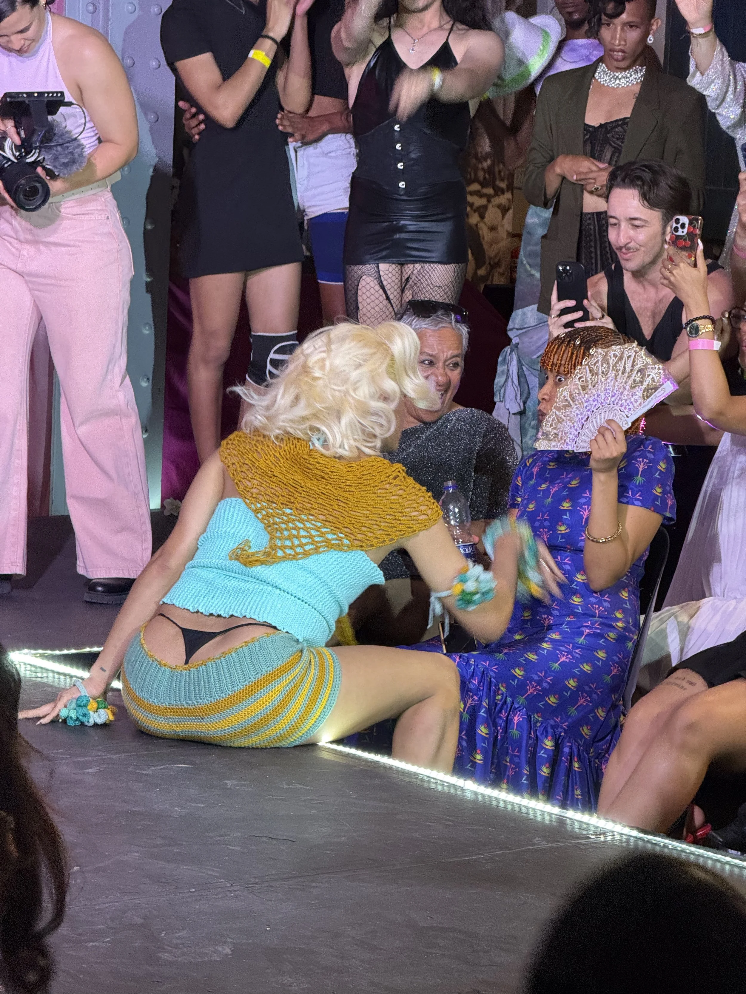 A scene at a lively event with two women sitting on the floor, one with platinum blonde hair wearing a crocheted yellow and blue outfit, and the other holding a folding fan and wearing a blue dress with colorful patterns. Several people are standing 