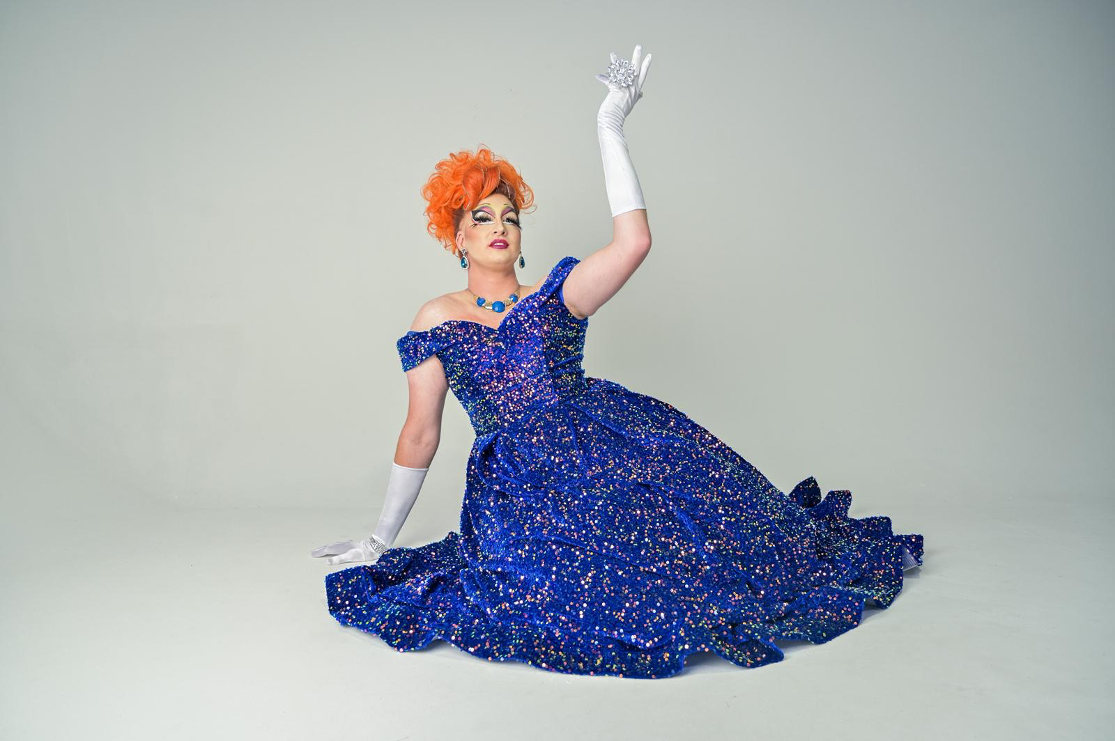 A drag queen with orange curly hair wearing a sparkly blue gown, white gloves, and jewelry, posing against a plain background.
