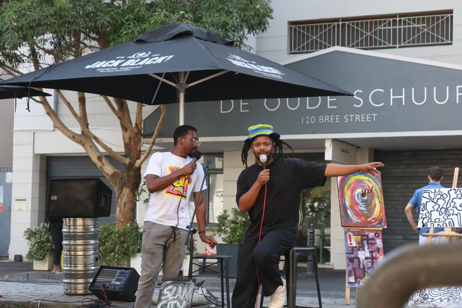 Two men are on a stage outdoors, one speaking into a microphone, the other listening. Behind them are paintings on easels, a large umbrella, and a sign that reads "DE OUDE SCHUUR 120 BREE STREET." One man is wearing glasses and a white t-shirt with colorful graphics, the other is wearing a black shirt and colorful hat with dreadlocks.