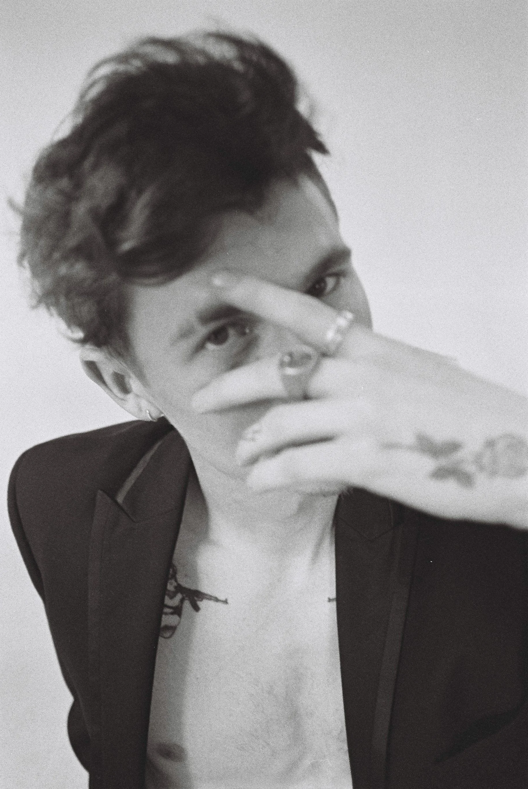 Black and white photo of a person with short, wavy hair wearing a dark blazer, holding their hand over their face, revealing tattoos on their neck and fingers.