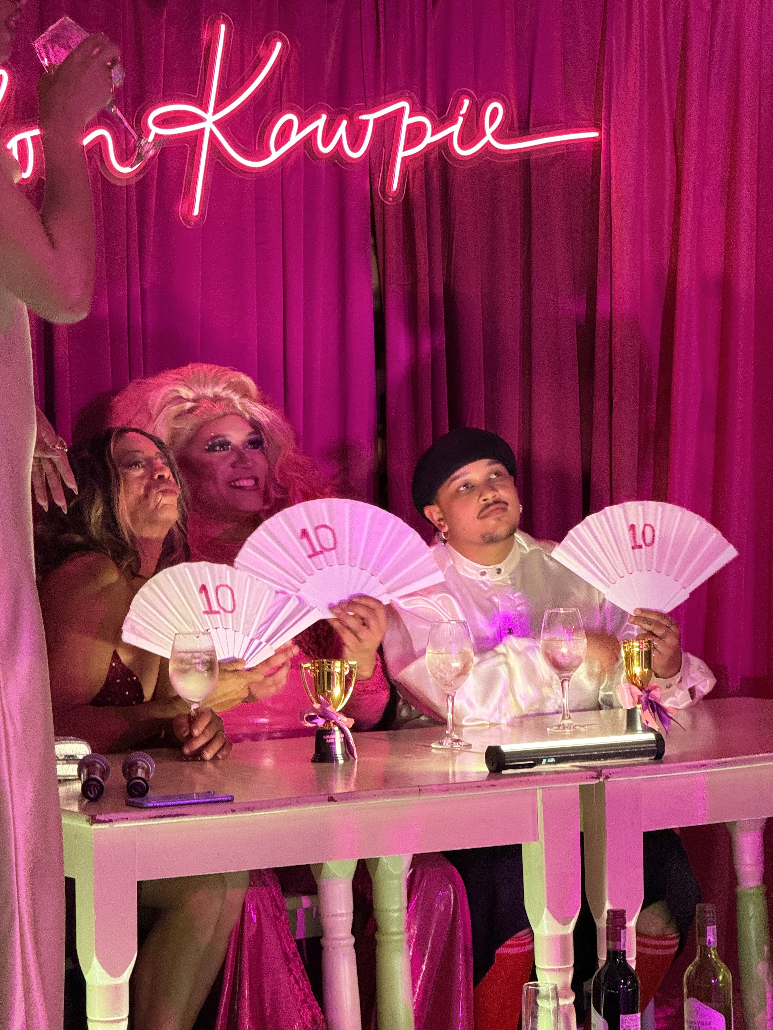 People sitting at a table with pink curtains and neon signs in the background, holding fans with the number 10 and trophies, in a pink-lit room.