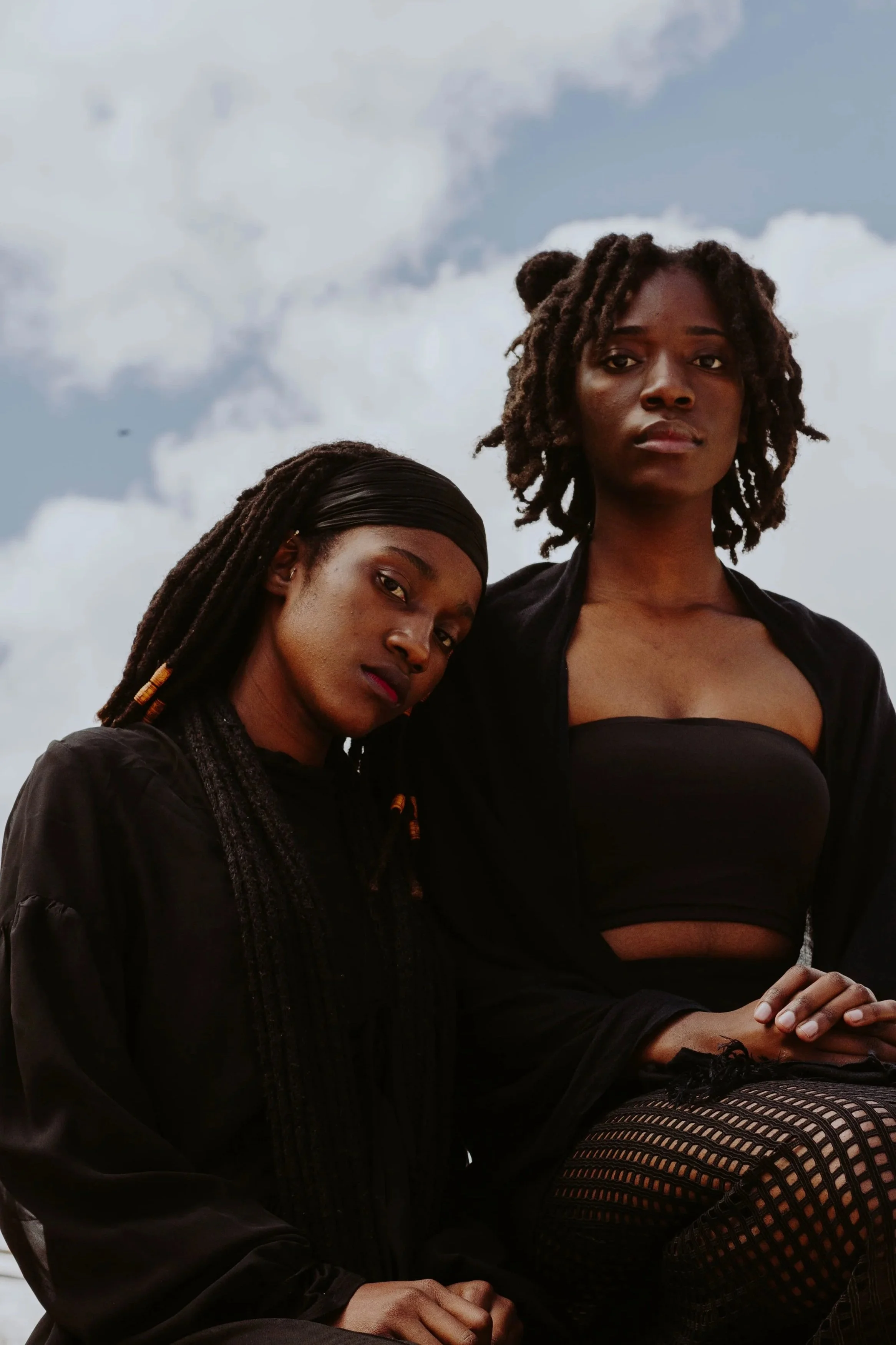 Two women with dark skin and curly hair posing against a cloudy sky, dressed in black clothing, one with natural dreadlocks and the other with an updo.