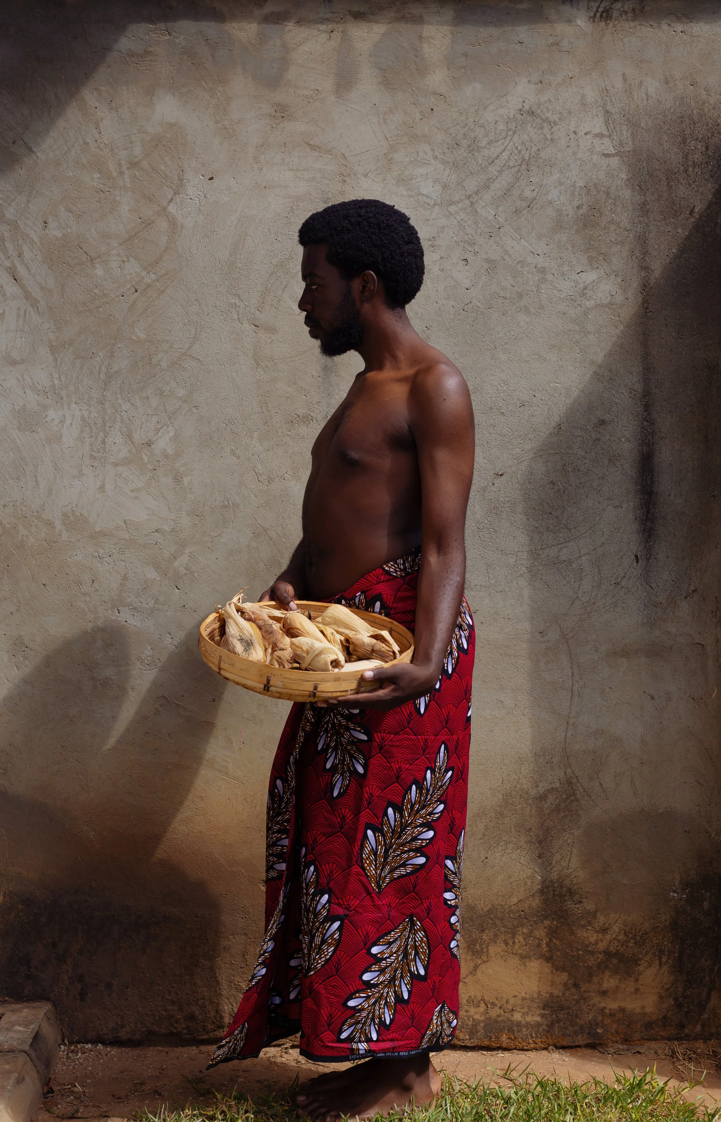 A shirtless man wearing a colorful patterned cloth around his waist holding a round basket of dried corn ears, standing barefoot against a textured beige wall with a shadow cast behind him.