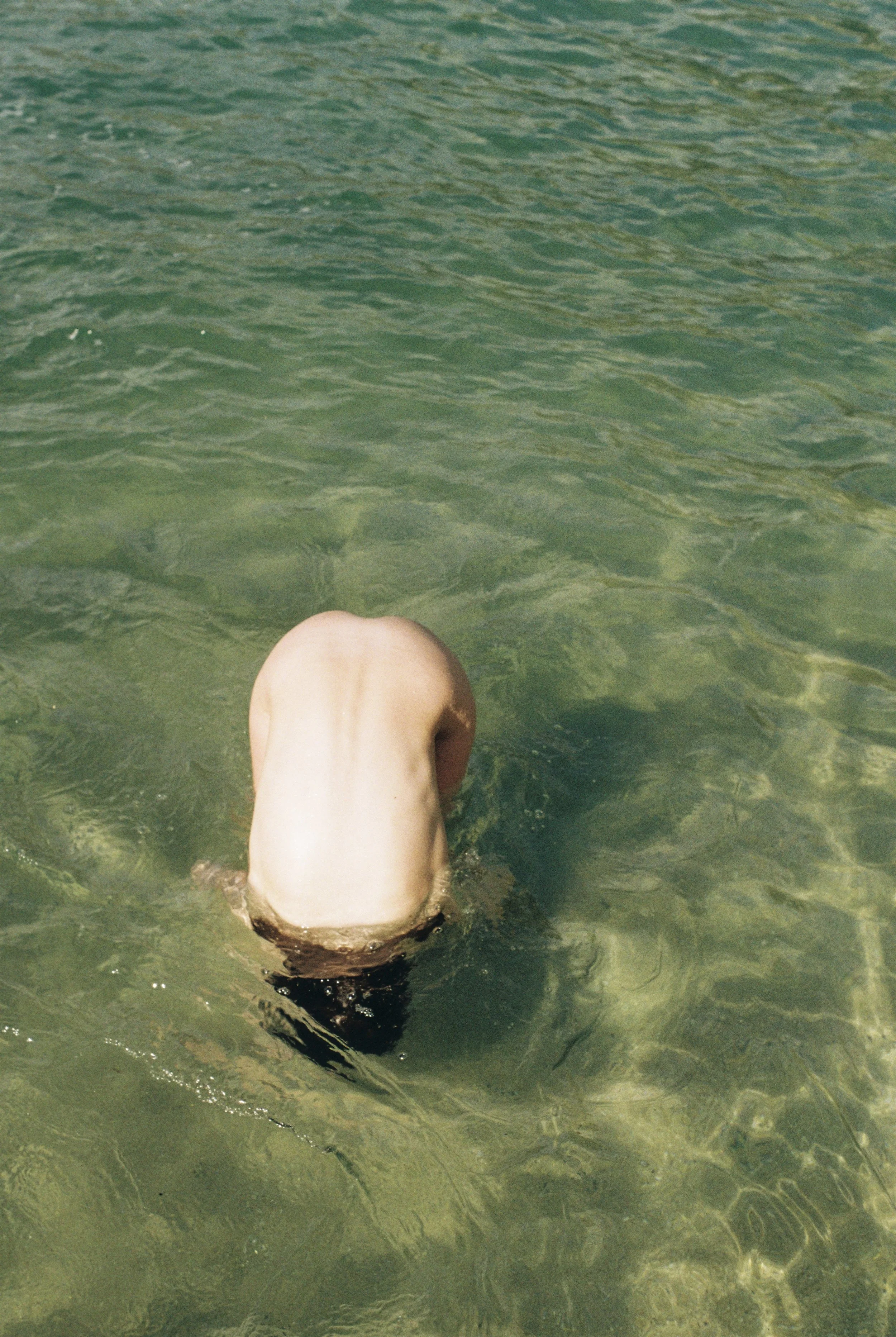 Person with light skin and short dark hair submerged in clear green water, with their head facing down.