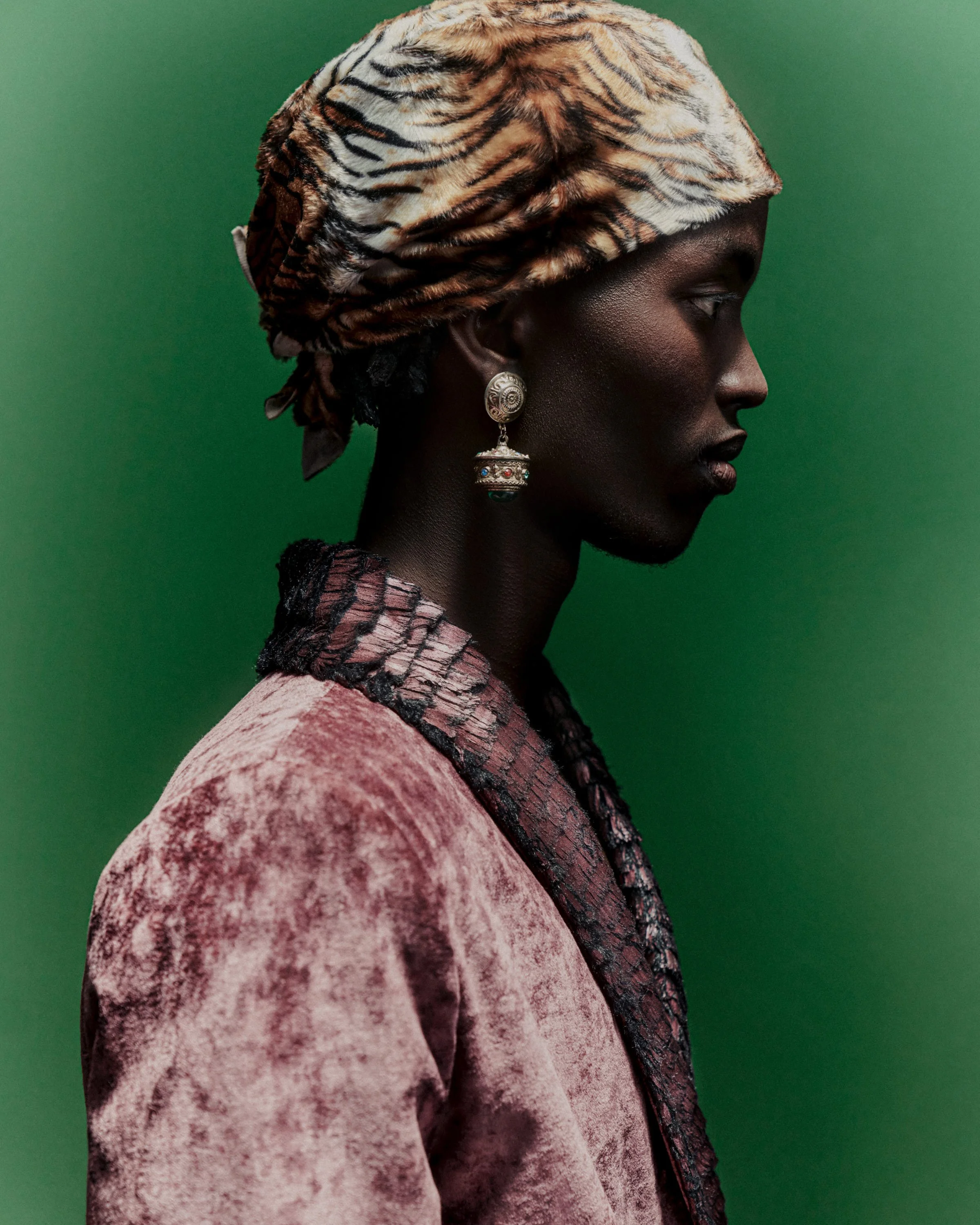 Profile of a woman with dark skin wearing a tiger-print headscarf, large earrings, and a textured pink and black outfit against a green background.