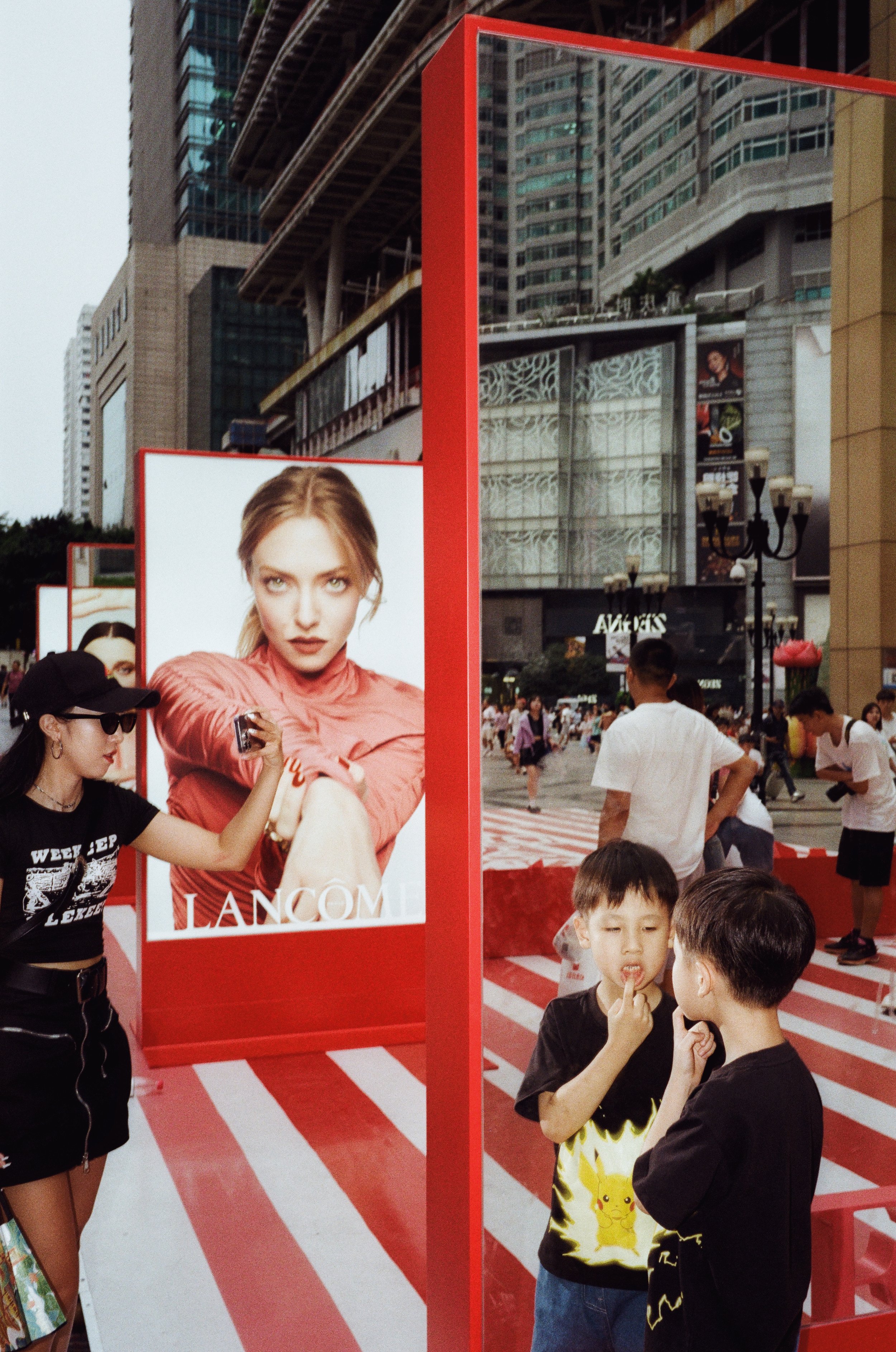 People at an outdoor event in an urban area with large advertisement posters, including a Lancôme ad featuring a woman with blonde hair in a pink top, high-rise buildings in the background, and children and adults walking around.