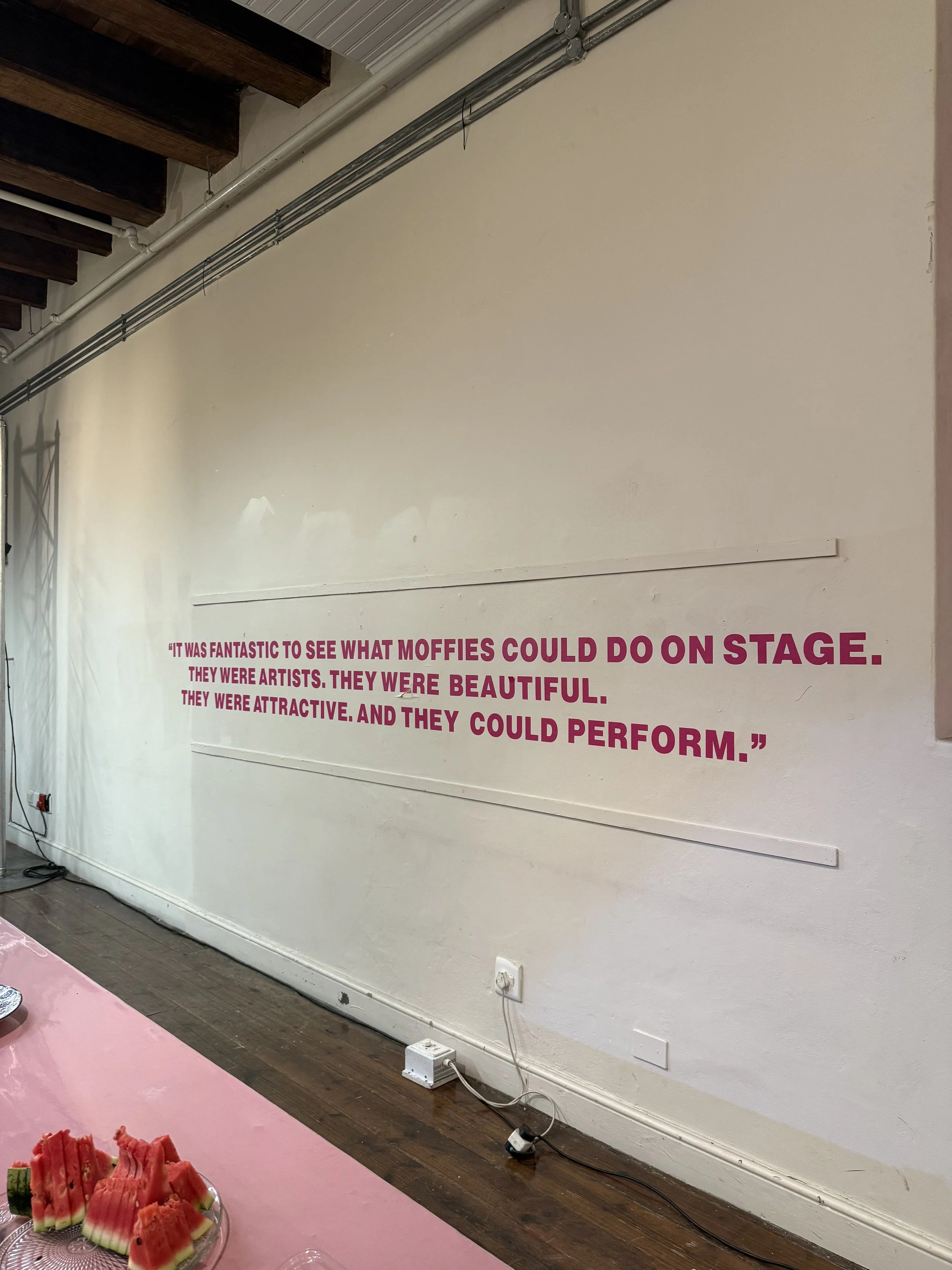 A white wall with a quote in large pink text that reads, 'It was fantastic to see what Moffies could do on stage. They were artists. They were beautiful. They were attractive, and they could perform.' There are some electrical outlets and cords near 
