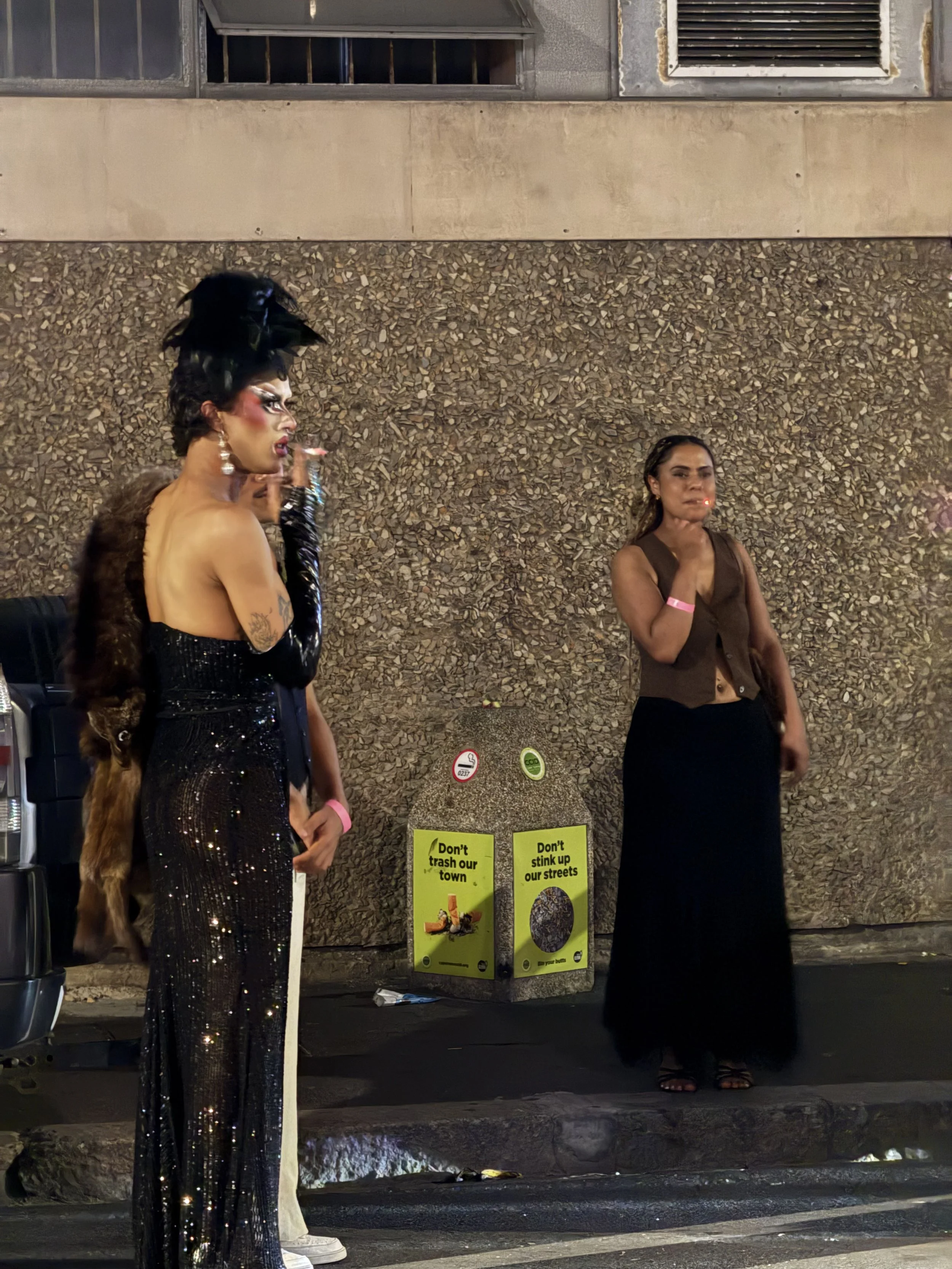 Two drag queens standing on the sidewalk in front of a textured wall at night. One is in a black sequined dress with a large black feathered headpiece, and the other in a brown vest and black skirt. A yellow trash bin with signs reading 'Don't trash 