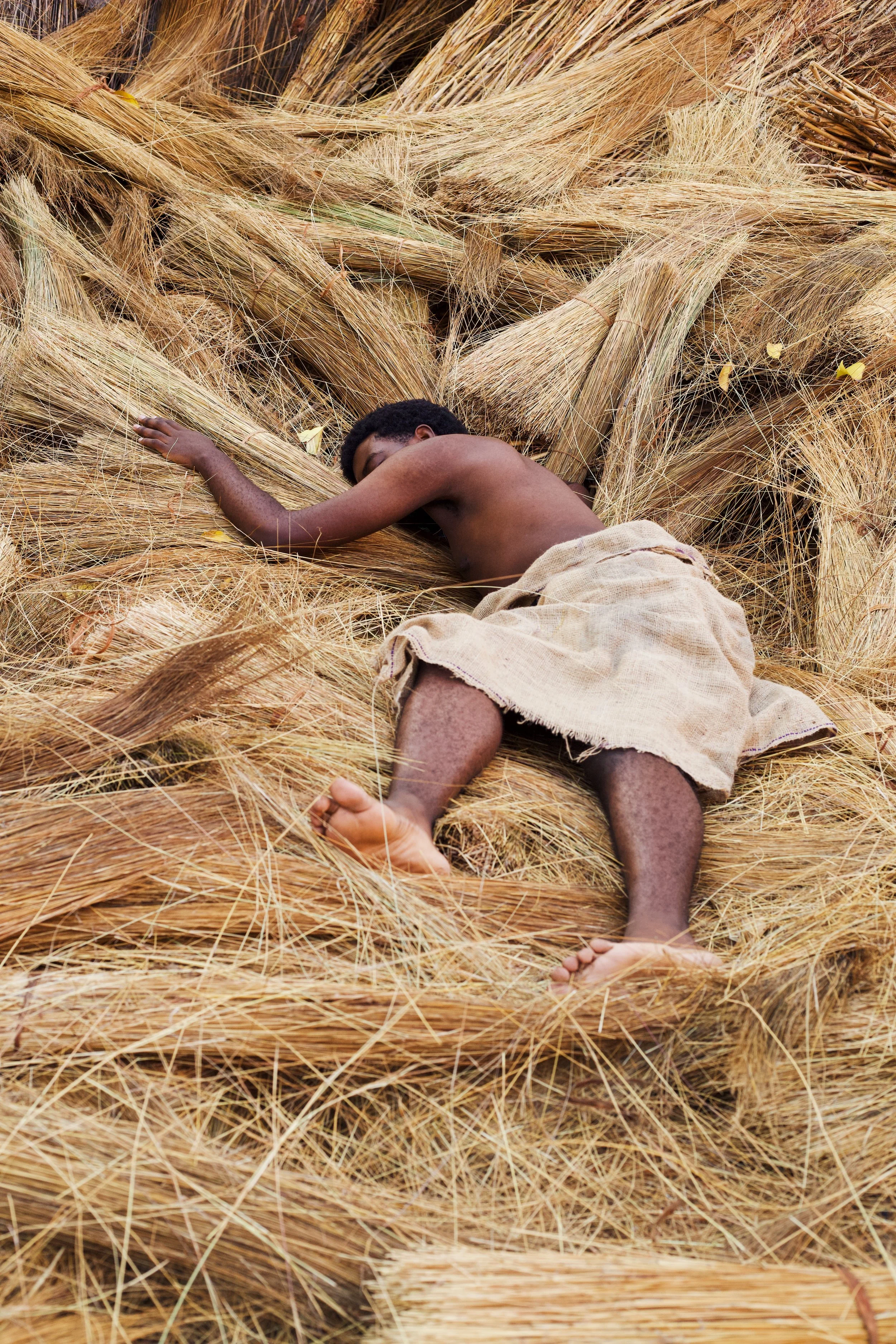 A young boy is lying on yellow straw, sleeping or resting with his arm extended and eyes closed, wearing a beige cloth around his waist.