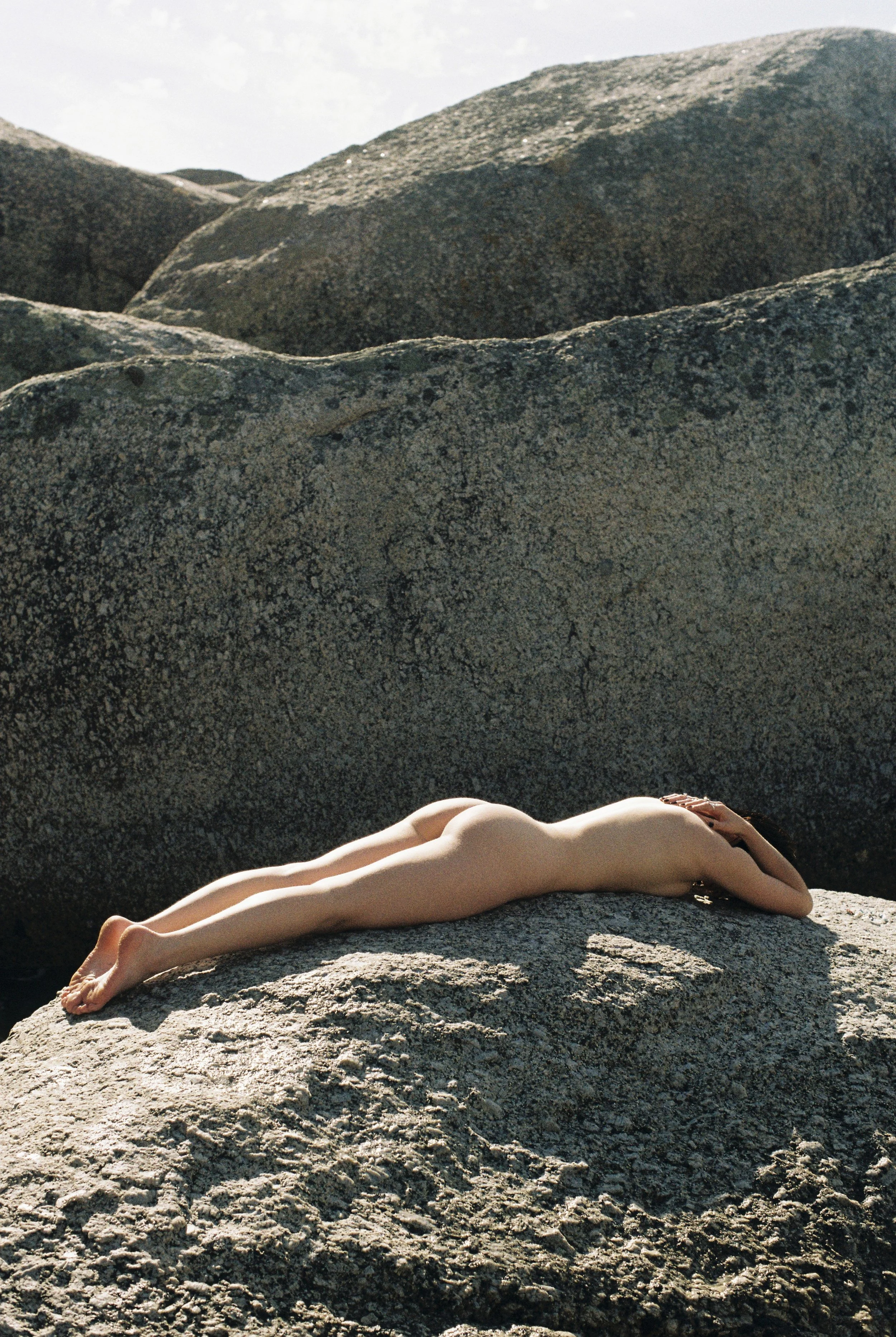 A nude woman lying face down on a large rock surrounded by boulders outdoors.