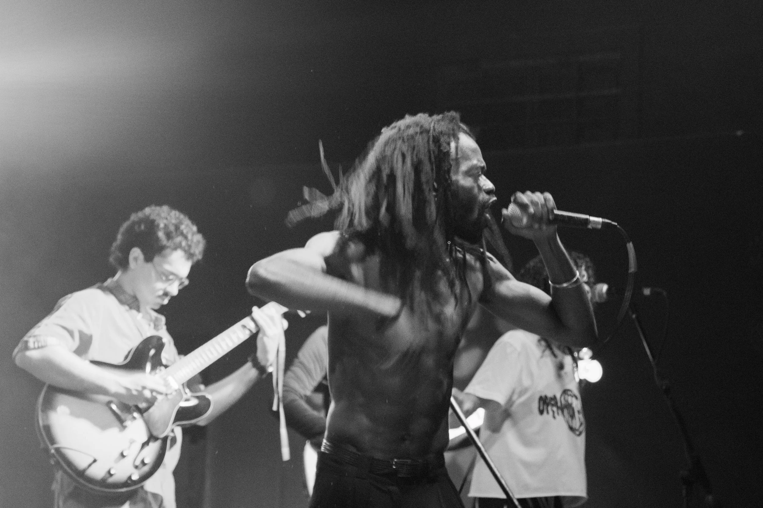 A shirtless man with dreadlocks passionately singing into a microphone during a live music performance, accompanied by a guitarist playing an electric guitar on a dimly lit stage.