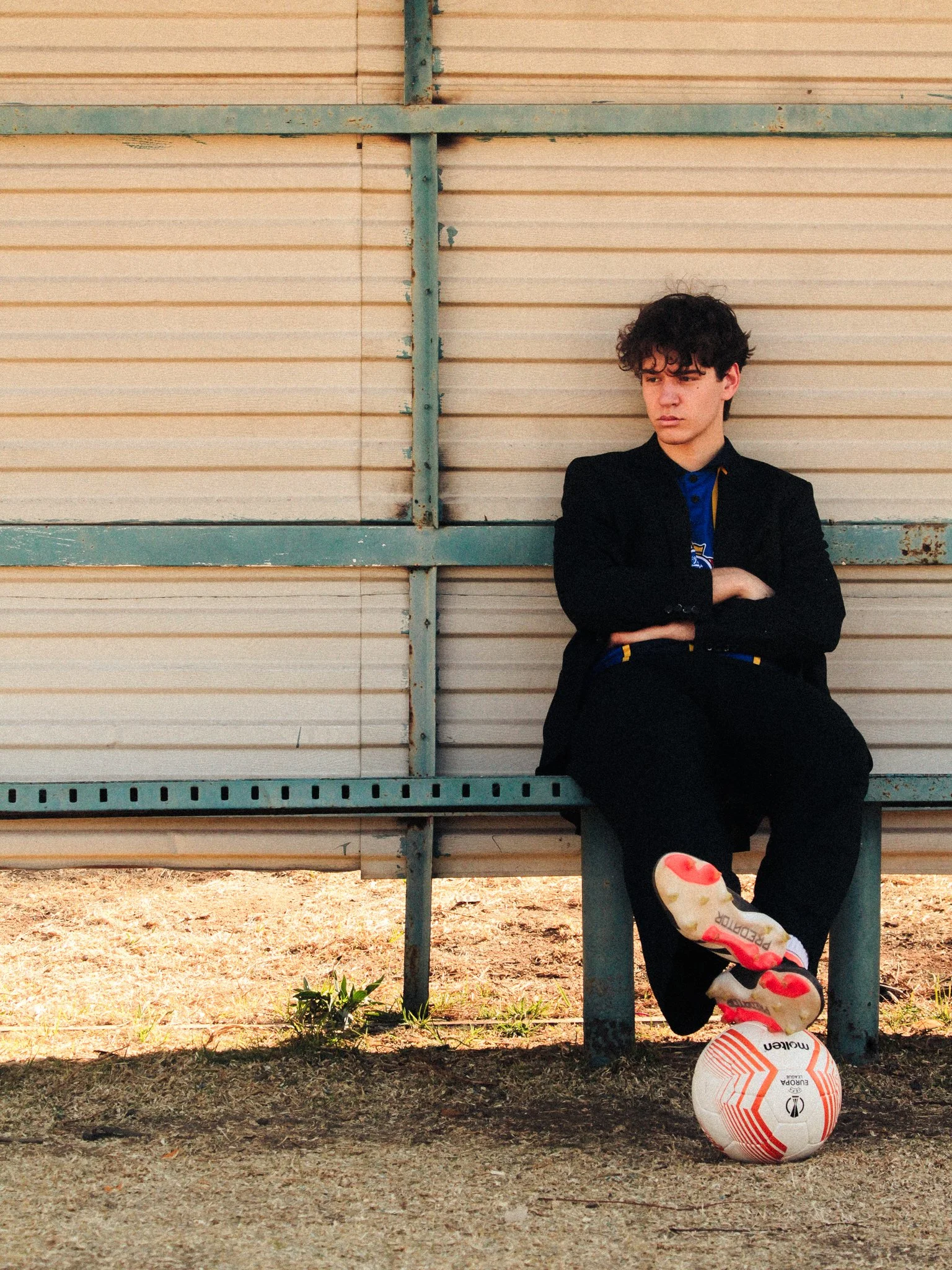 A young man in a black suit sitting on a bench with his arms crossed, looking to the side. A soccer ball is on the ground near his foot, which is resting on top of it. The background is a beige industrial wall.