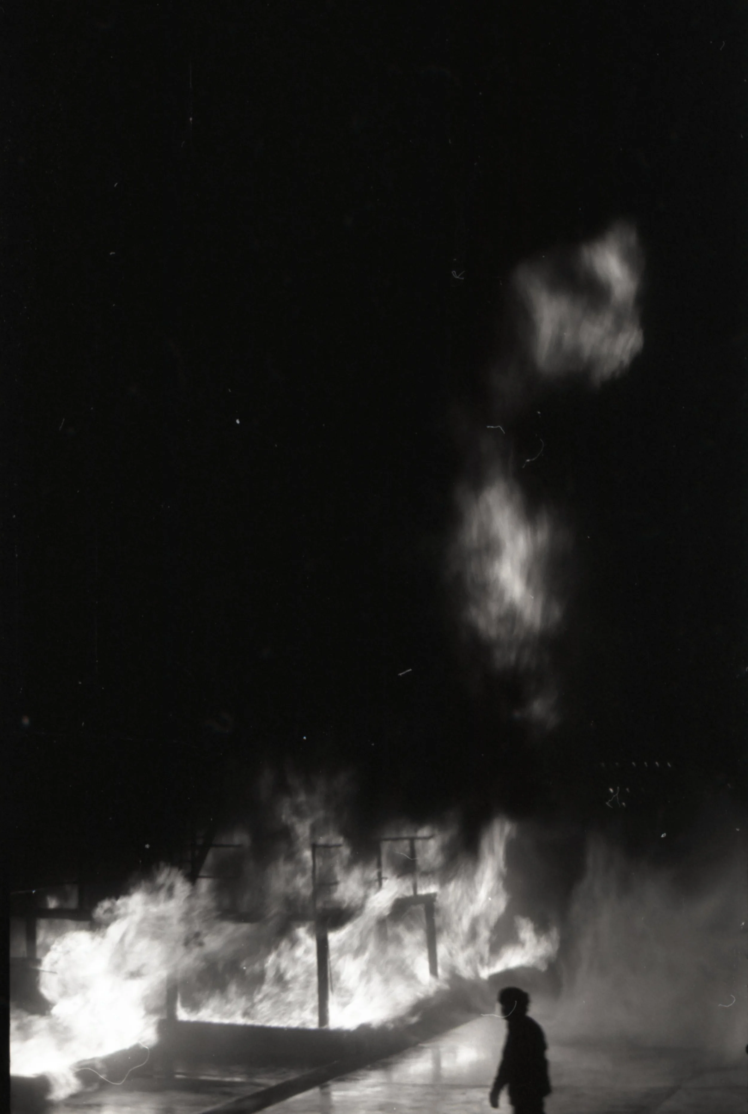 A person walking near a fire on a street at night, with large flames and smoke rising into the sky.