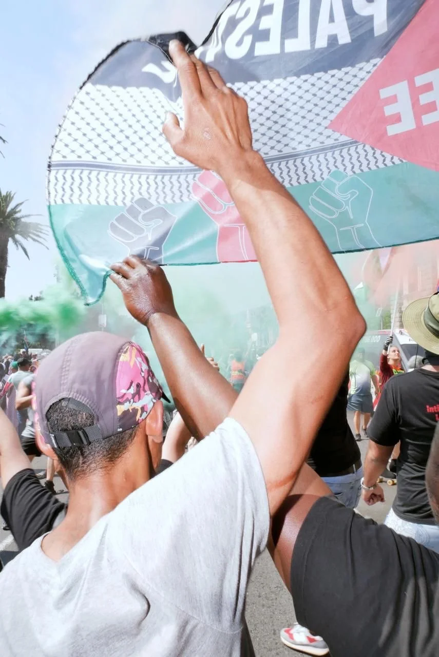 People holding up a large rainbow-colored banner or flag at a street protest or march, with a crowd in the background and some green smoke in the air.