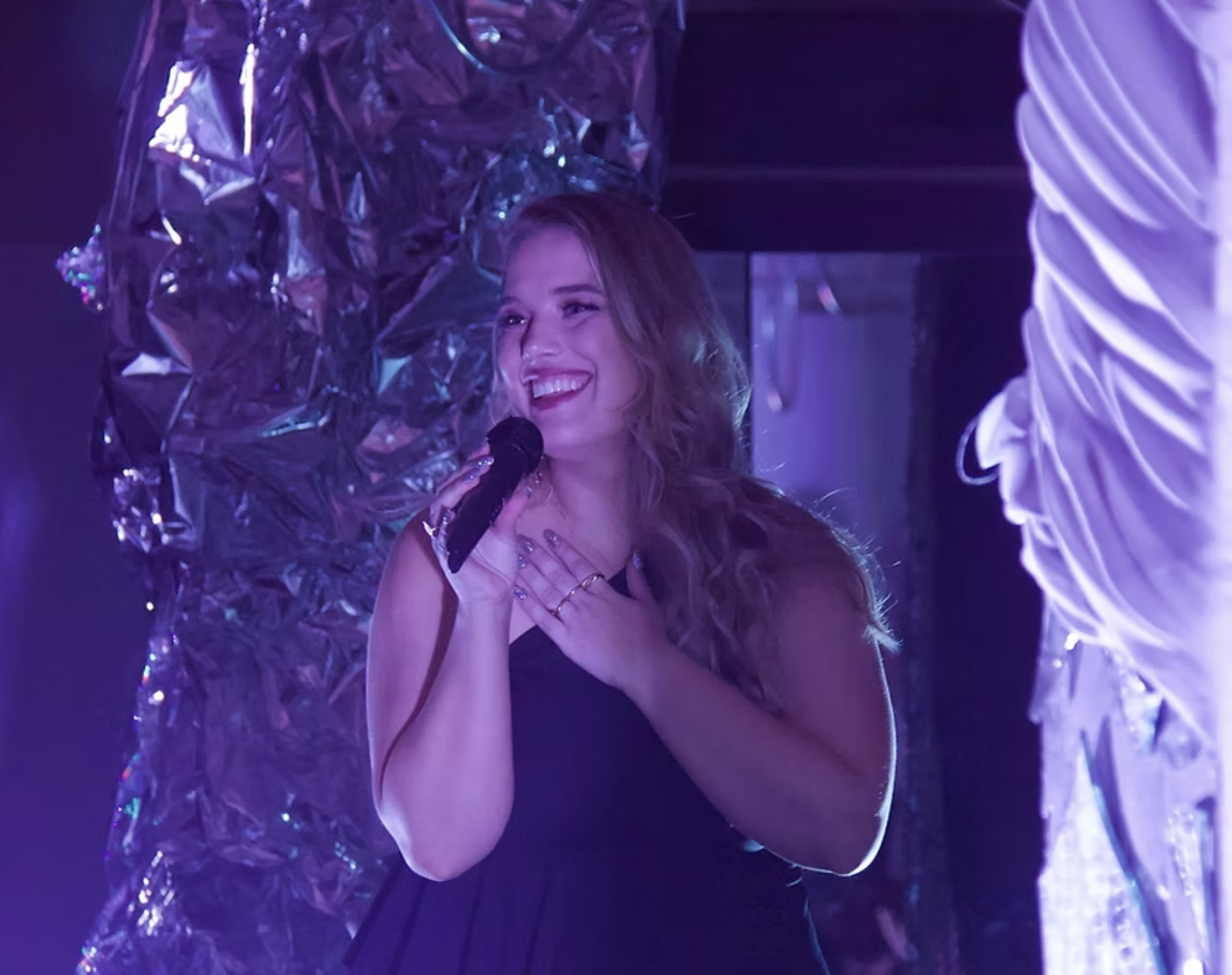 Young woman with curly hair smiling and holding a microphone, standing on stage with purple and silver decor in the background.