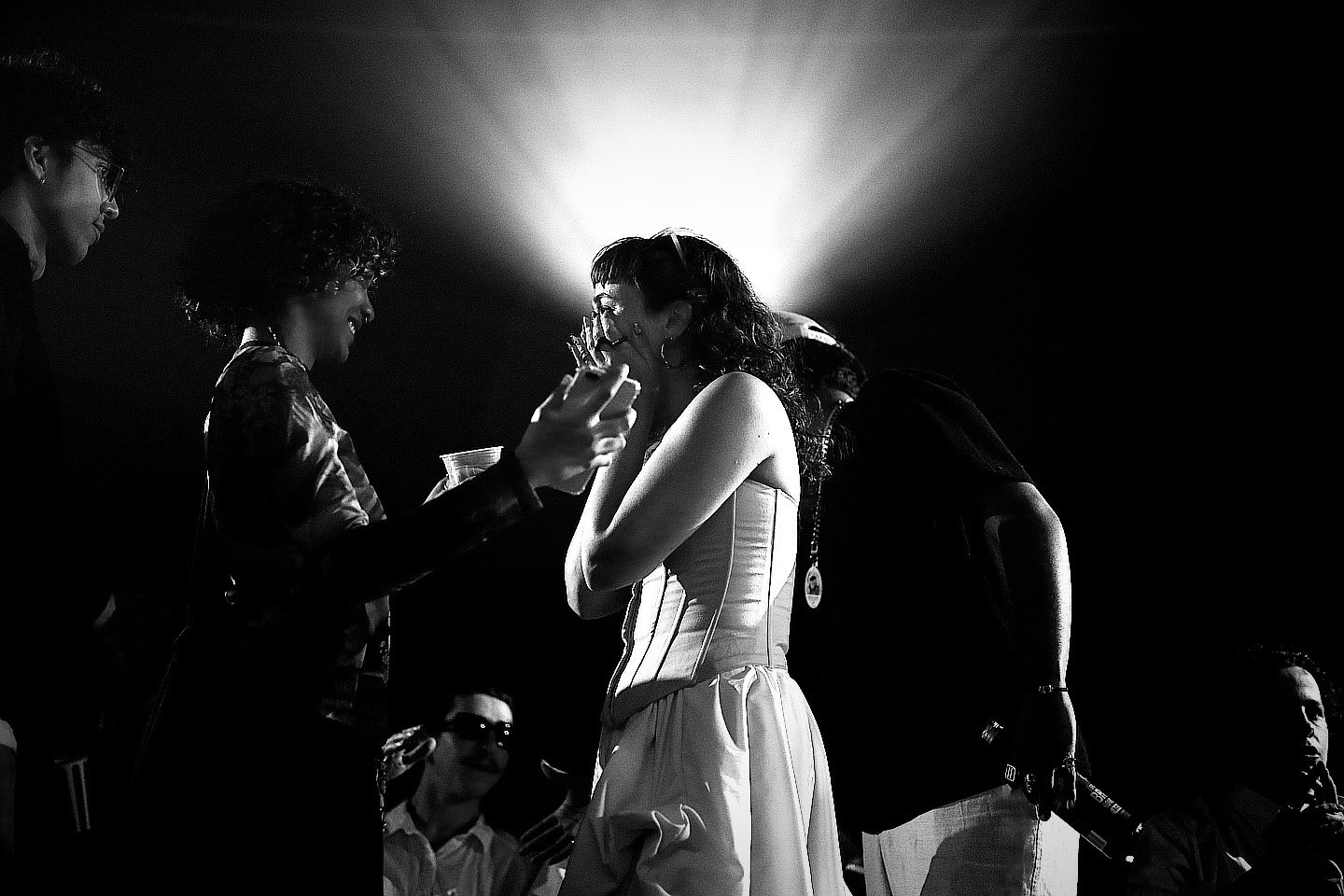 Black and white photo of a woman in a strapless dress smiling and talking to two women, one with curly hair and another with glasses, while they laugh.