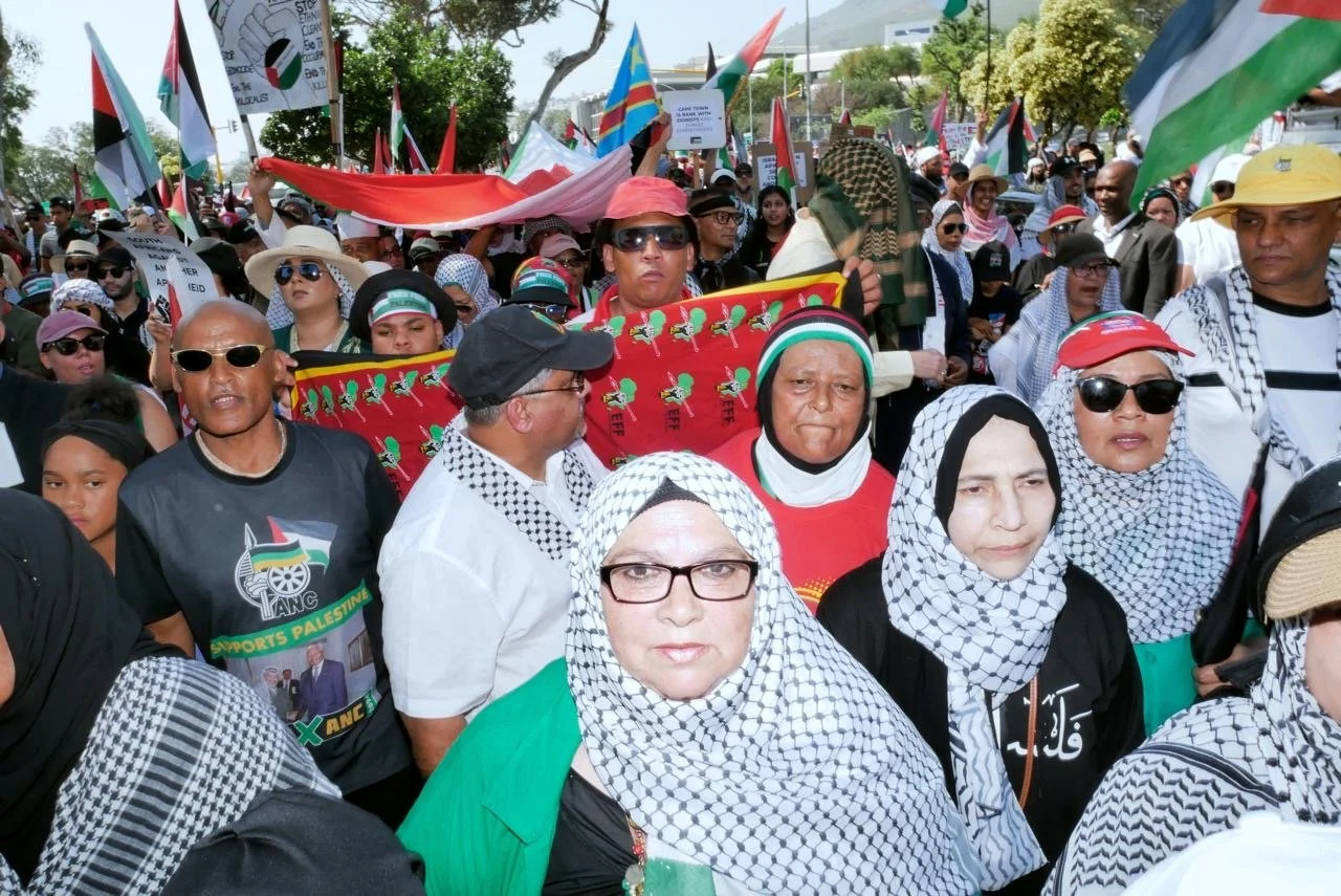A large crowd participating in a protest or rally, with many holding flags and signs related to Palestine. The crowd includes diverse men and women, some wearing keffiyeh scarves and patriotic clothing.
