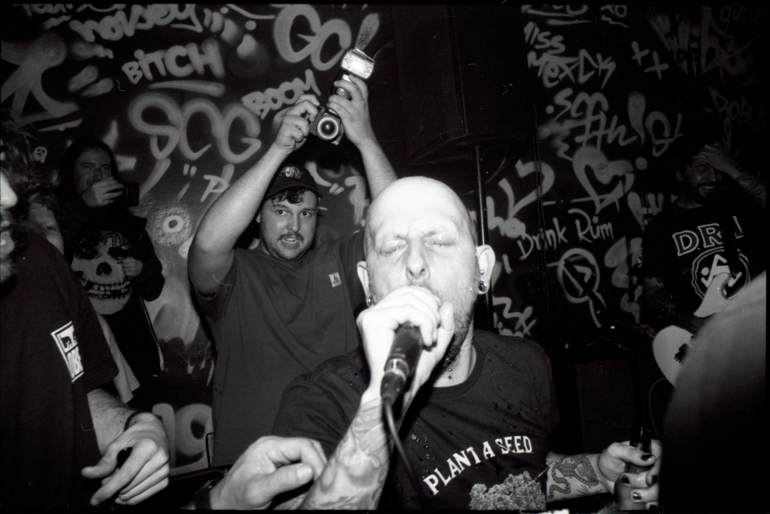 A musician passionately performing into a microphone with a clenched fist, surrounded by people at a concert or club, with graffiti-covered walls in the background.