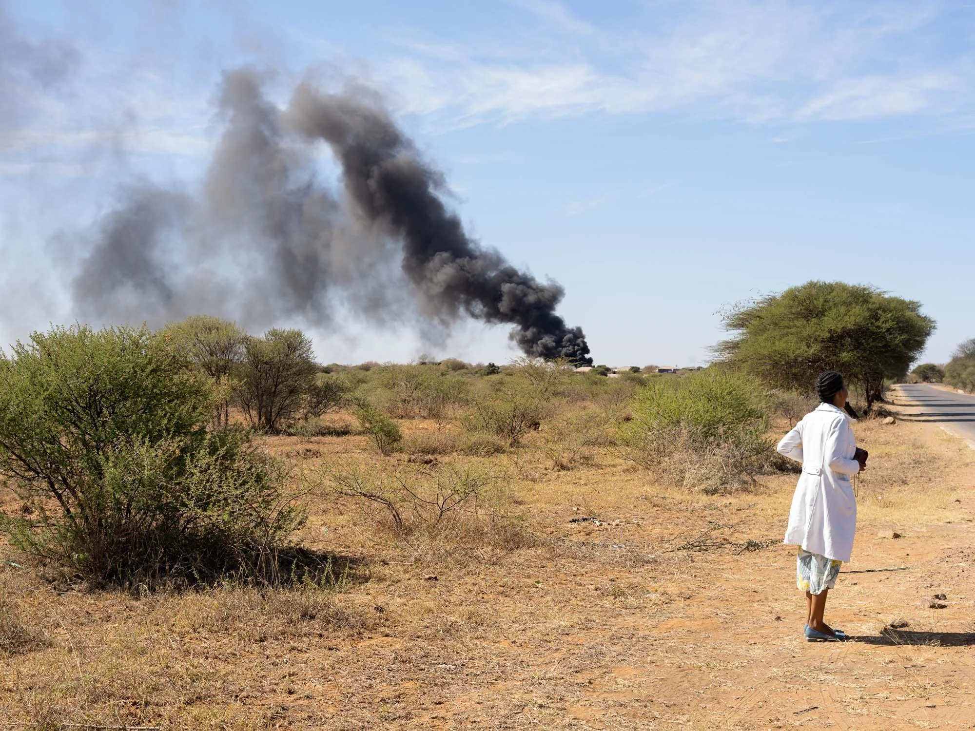 A woman in a white lab coat standing on a dirt road, observing a large plume of black smoke rising in the distance in a dry, sparsely vegetated landscape.