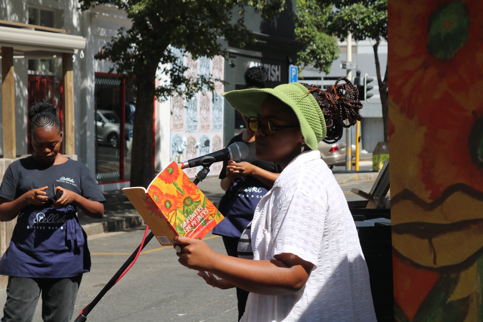 A woman with a green sun hat, glasses, and a white shirt is reading a colorful book into a microphone outdoors. There are two other women nearby, one looking at her phone. Trees, cars, and a city street are visible in the background.