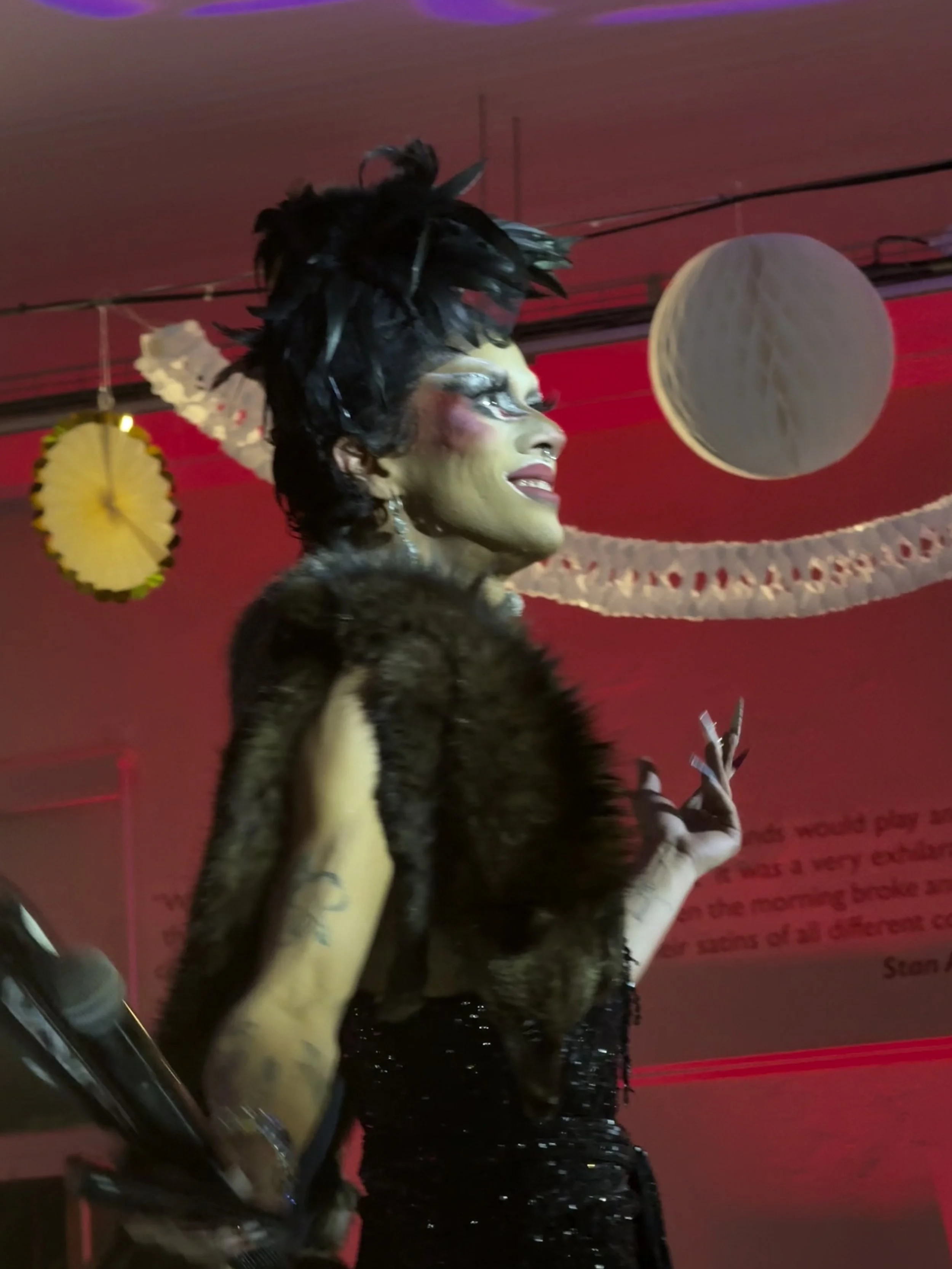 Person dressed in dramatic black costume with feathered headpiece, makeup, and long painted nails, standing in a decorated room with red lighting.
