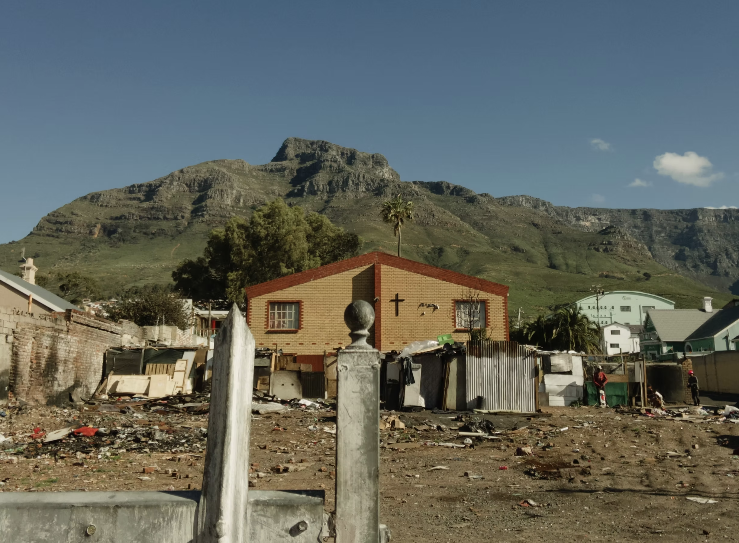 A rural area with a mountain in the background and a small church with a cross on the front in the foreground. There is debris and makeshift structures on the ground.
