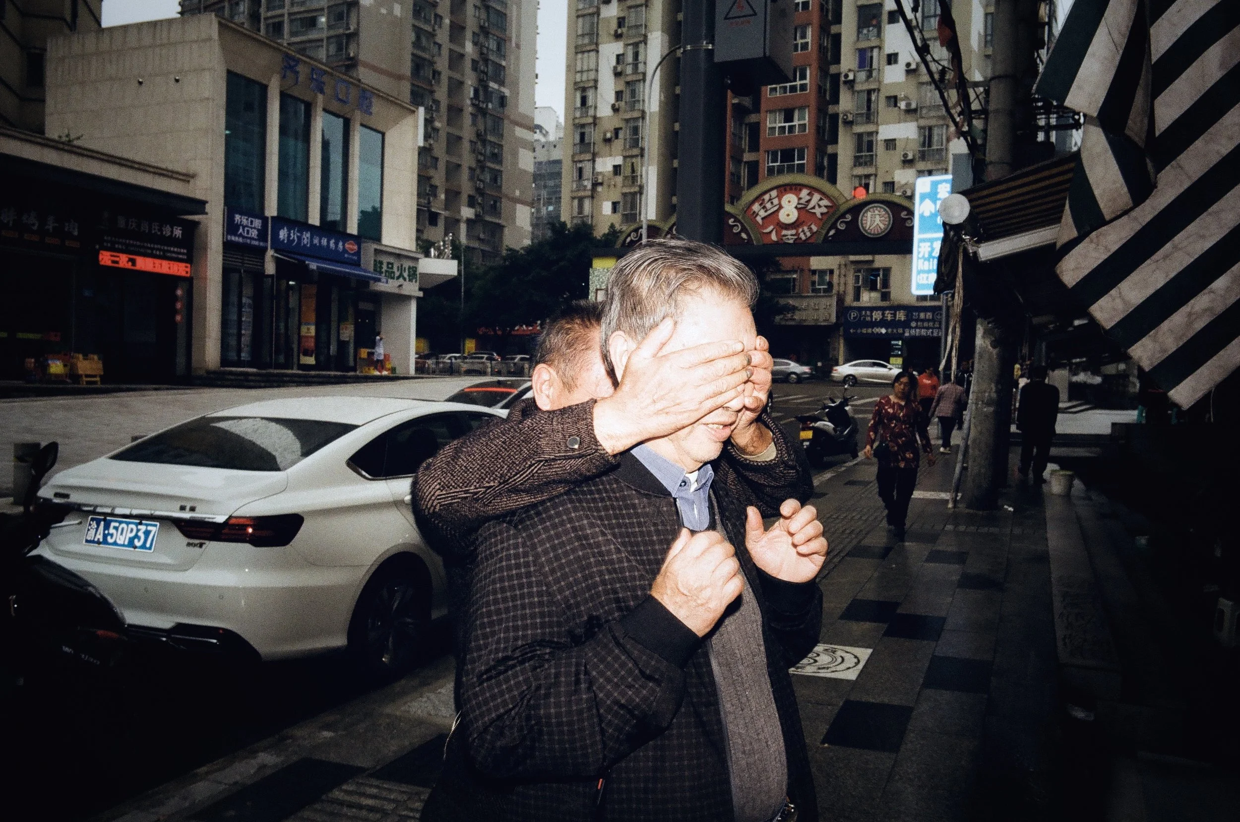A man stands on a city sidewalk with his eyes covered by another person's hands, in an urban setting with cars, pedestrians, and tall buildings.