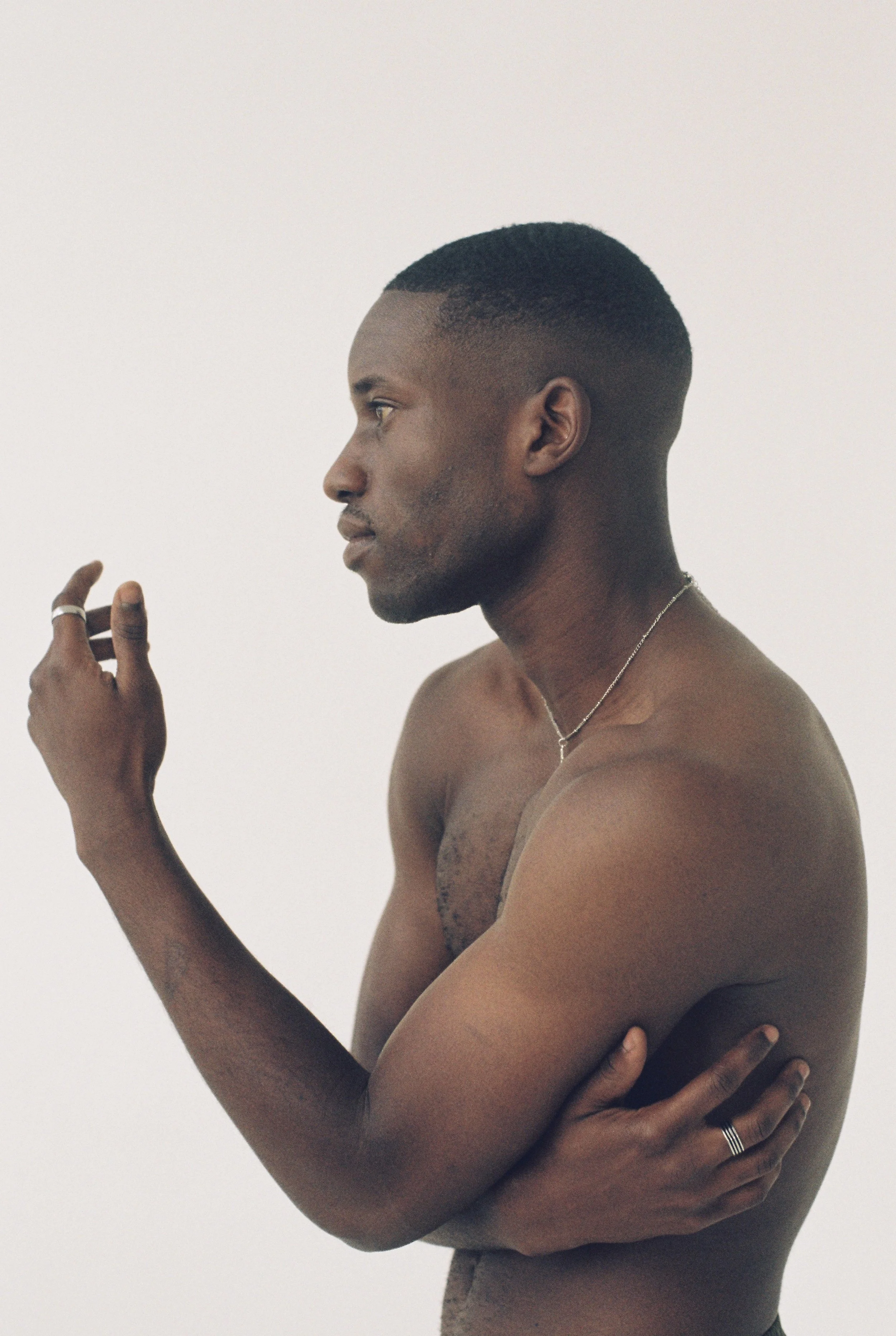 A shirtless man with short hair and jewelry stands against a plain white background, posed in profile with his right hand near his face and his left arm crossed over his chest.