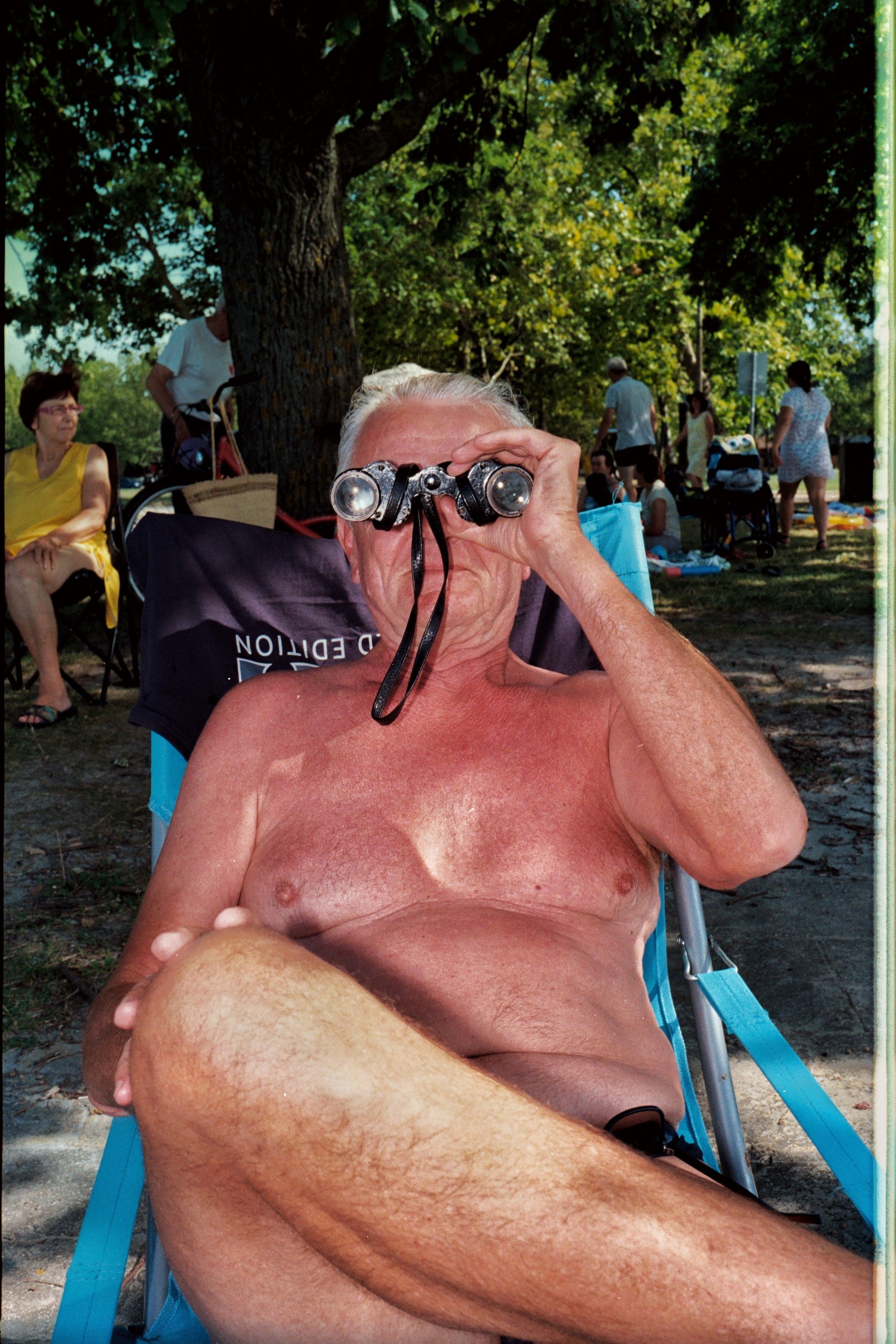 An elderly man with sunburned skin sitting outdoors under a tree, using binoculars, with people relaxing and standing in the background.